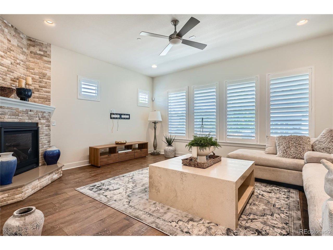 Image 2: 18x16 sq ft Family Room with stone-wall fireplace, wood floors, plantation shutters, and ceiling fan with a light. Image 2: 18x16 sq ft Family Room with stone-wall fireplace, wood floors, plantation shutters, and ceiling fan with a light.
