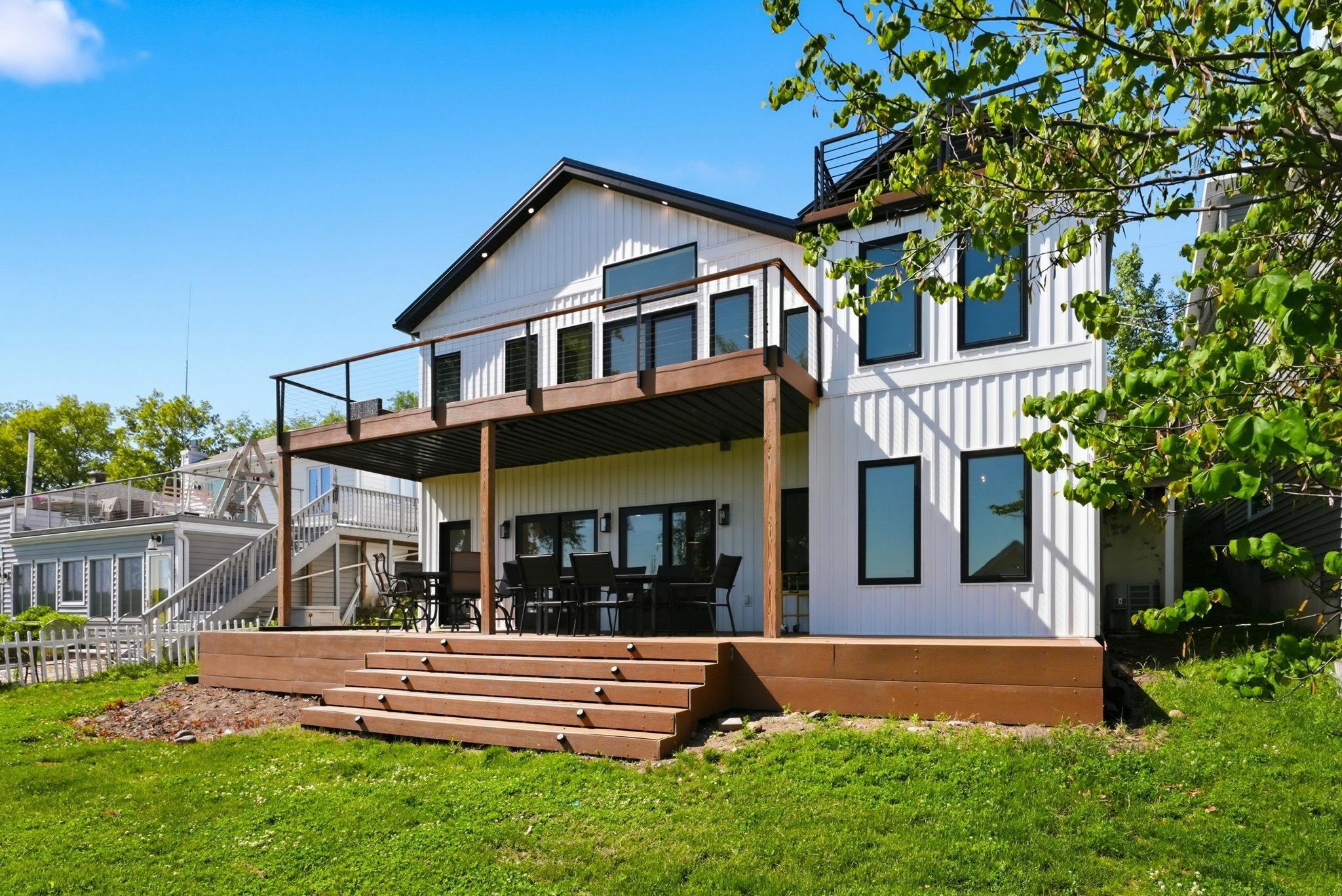 Image 2: Modern exterior featuring white vertical siding, black trim windows, a ground-level deck with integrated steps, and an upper-level balcony with glass railing, Back Of Structure Image 2: Modern exterior featuring white vertical siding, black trim windows, a ground-level deck with integrated steps, and an upper-level balcony with glass railing, Back Of Structure