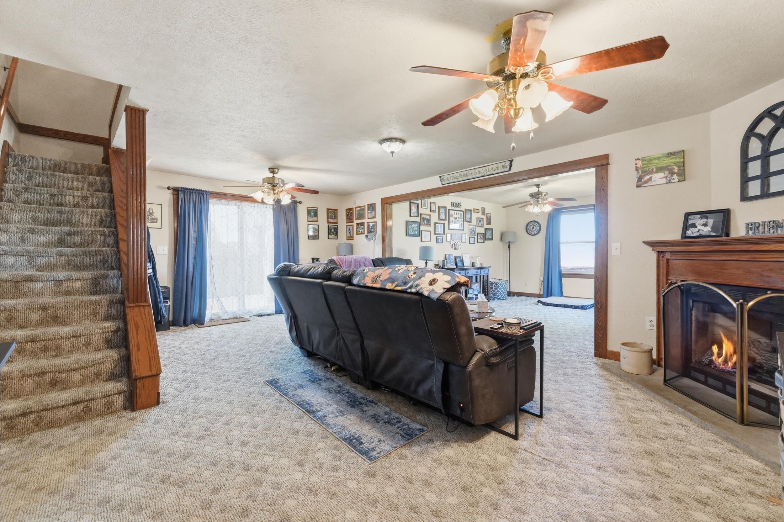 Image 2: Spacious living area featuring a fireplace with a wood mantle, carpet flooring, a ceiling fan, and a sliding glass door