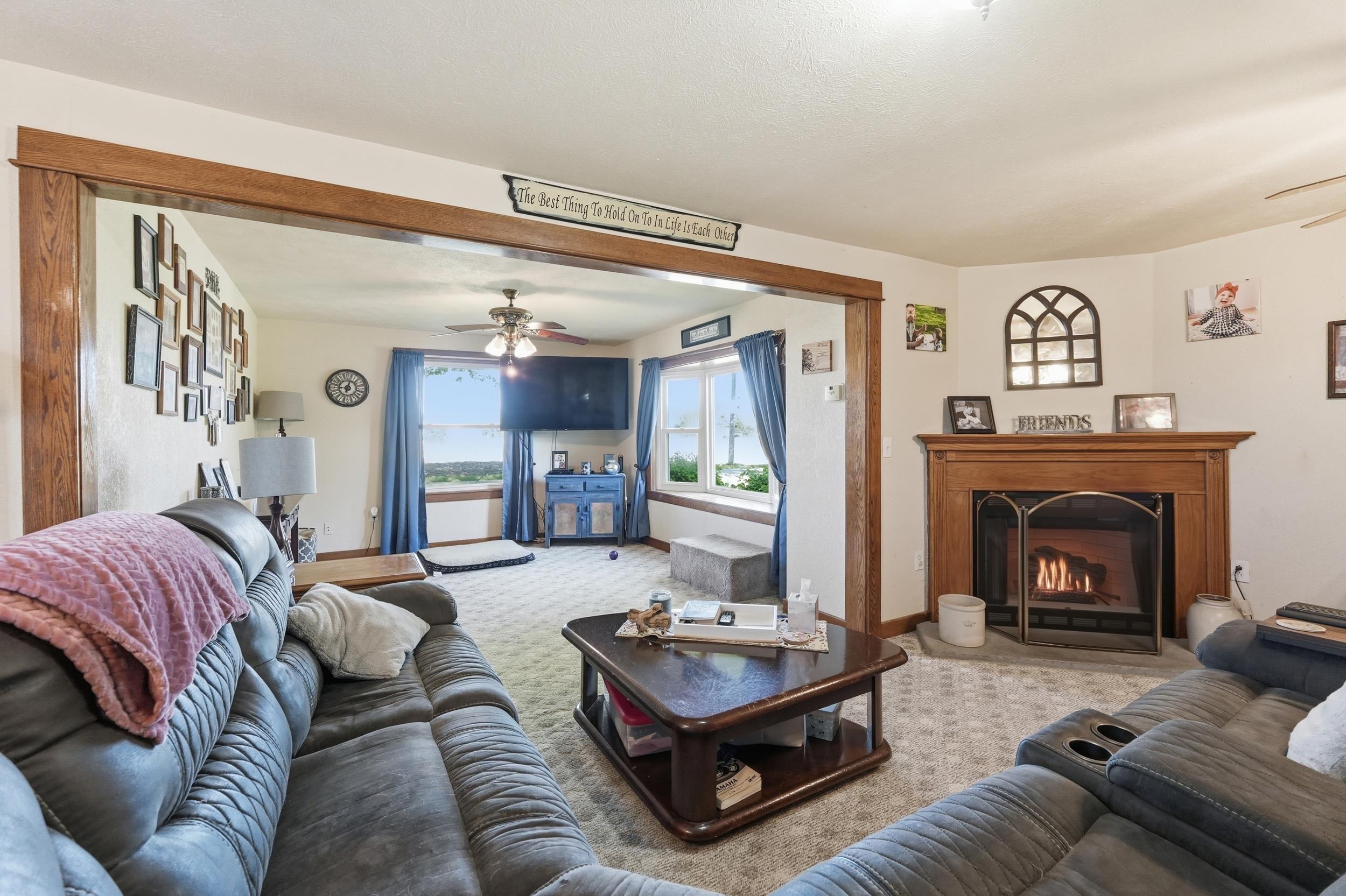 Image 3: Living area featuring a corner fireplace with a wood mantel, a ceiling fan, and large windows with exterior visibility