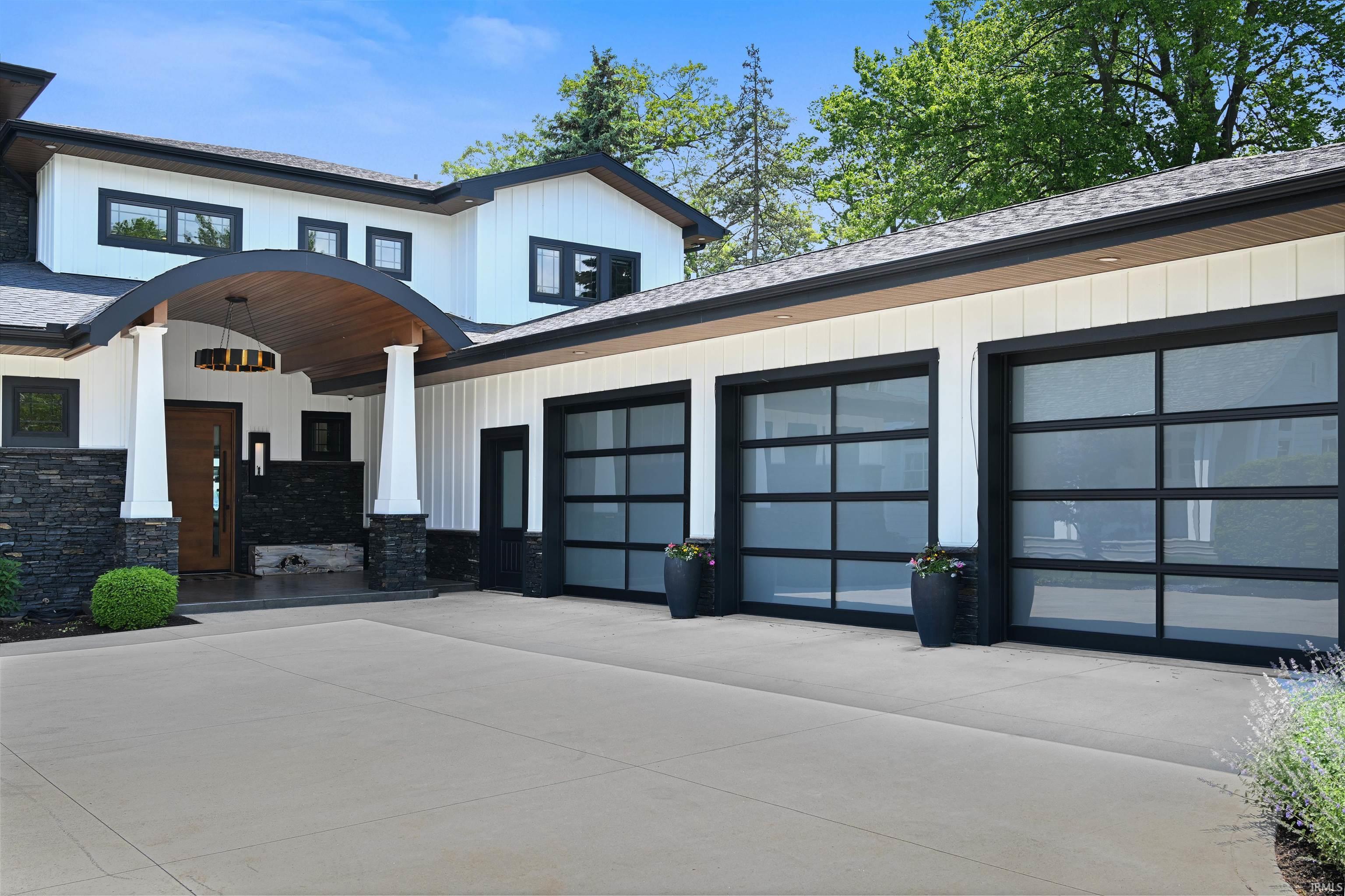 Image 3: Contemporary white exterior with black trim, three frosted glass garage doors, a curved wood-finish entry canopy, black stone accents, and a light-colored driveway, Front Of Structure