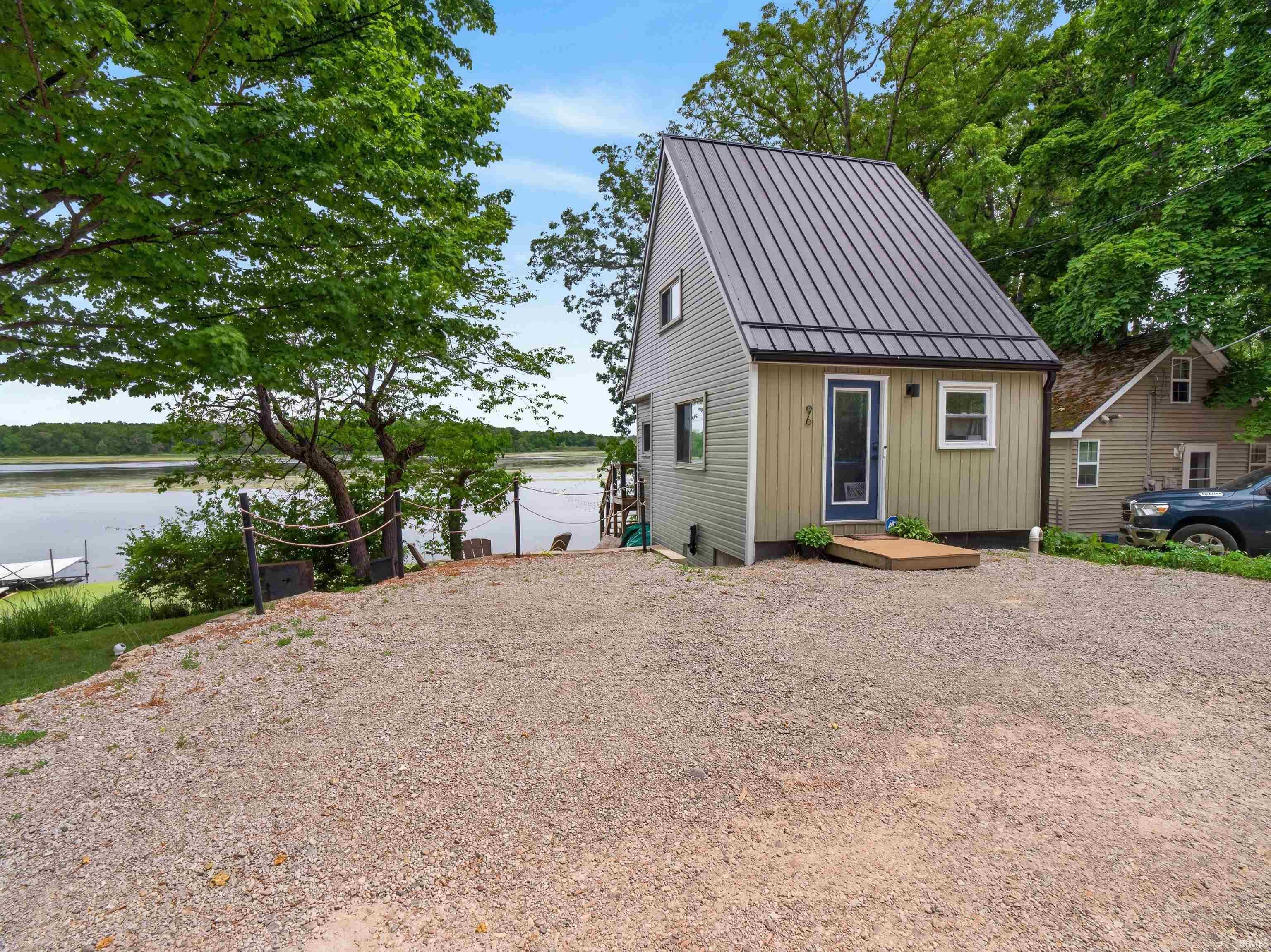Image 0: Waterfront property featuring a contemporary A-frame structure with metal roofing, vertical and horizontal siding, a gravel driveway, and mature landscaping, Front Of Structure Image 0: Waterfront property featuring a contemporary A-frame structure with metal roofing, vertical and horizontal siding, a gravel driveway, and mature landscaping, Front Of Structure