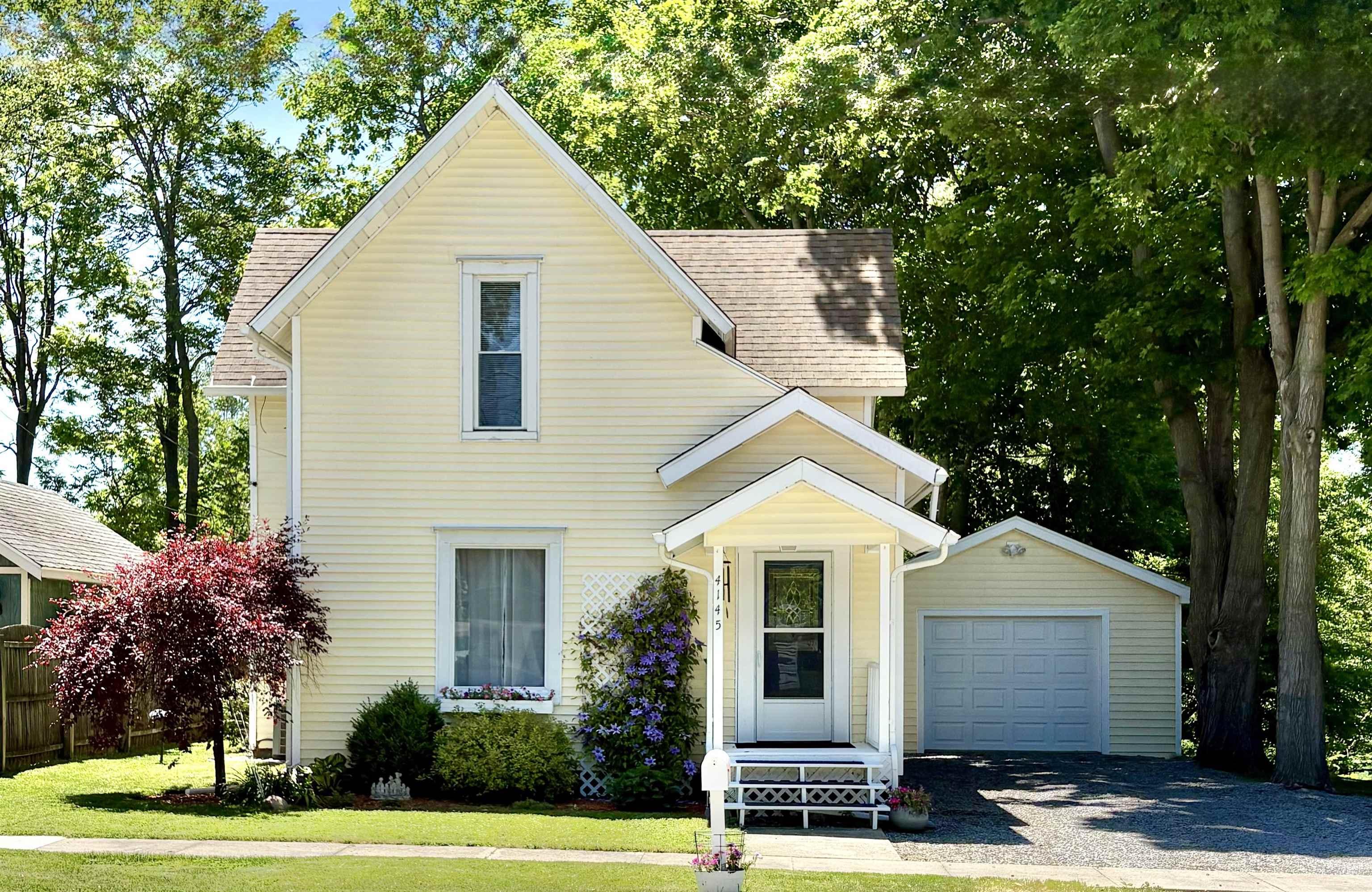 Image 0: Light yellow siding exterior with a peaked roofline, featuring a covered front porch with white railings, an attached single-car garage, and established front yard landscaping, Front Of Structure Image 0: Light yellow siding exterior with a peaked roofline, featuring a covered front porch with white railings, an attached single-car garage, and established front yard landscaping, Front Of Structure