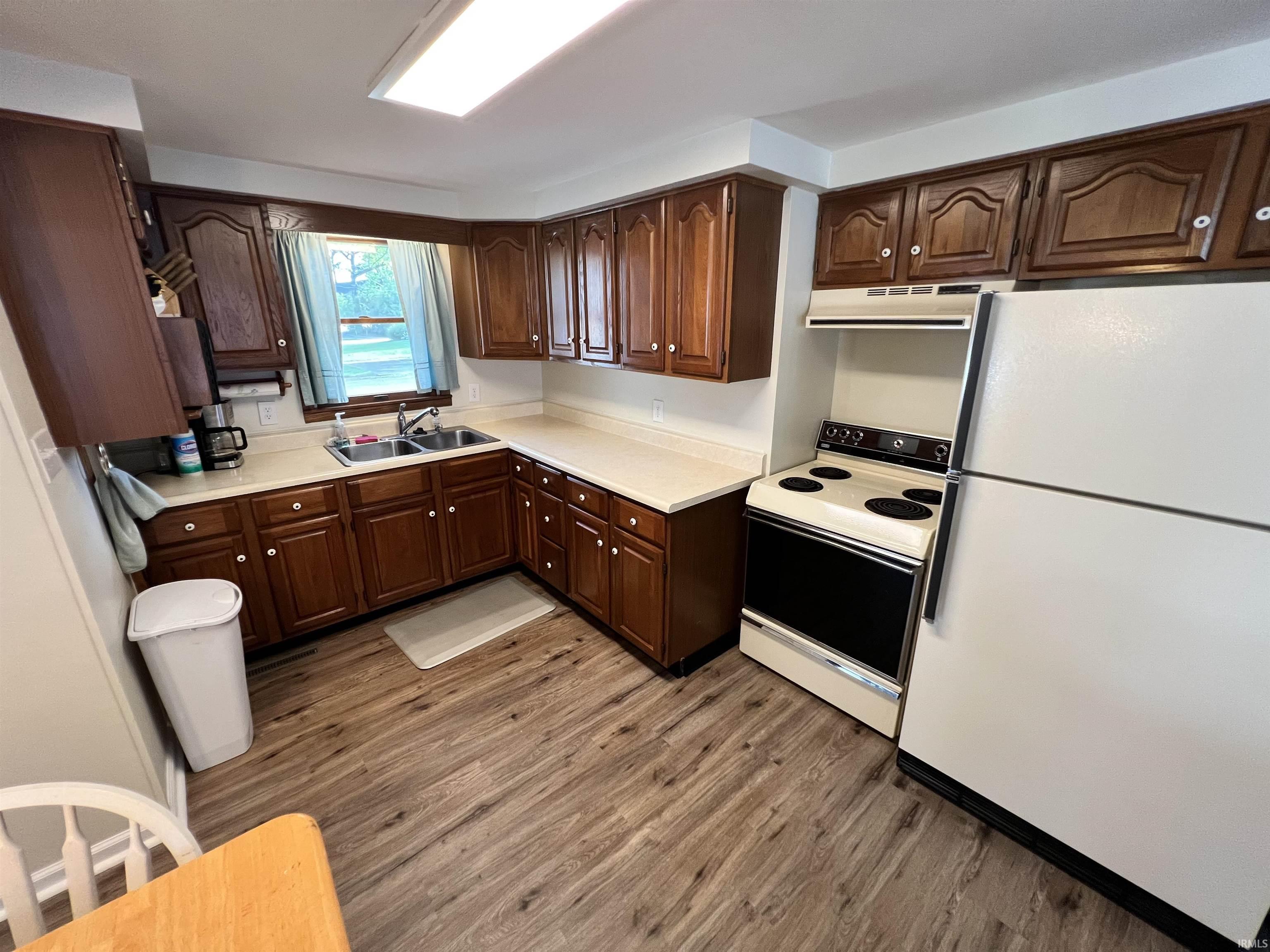 Image 1: Kitchen featuring wood-finish flooring, dark wood cabinetry, light countertops, and a double basin sink beneath a window, Kitchen Image 1: Kitchen featuring wood-finish flooring, dark wood cabinetry, light countertops, and a double basin sink beneath a window, Kitchen