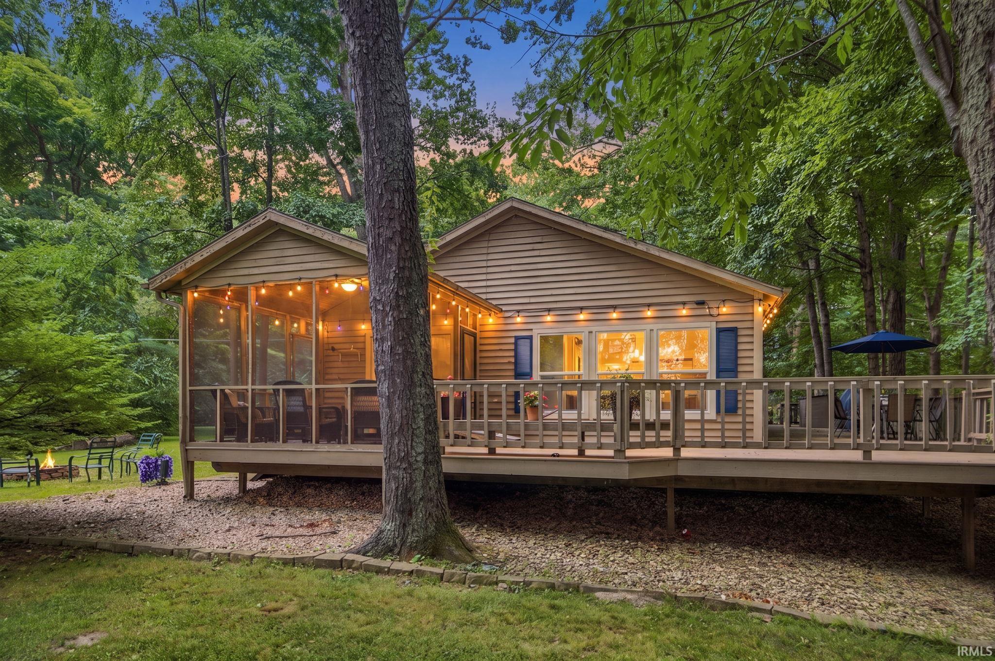 Image 1: Wood-sided exterior featuring a screened porch, expansive outdoor deck, multiple windows with blue shutters, and a surrounding wooded landscape, Back Of Structure Image 1: Wood-sided exterior featuring a screened porch, expansive outdoor deck, multiple windows with blue shutters, and a surrounding wooded landscape, Back Of Structure