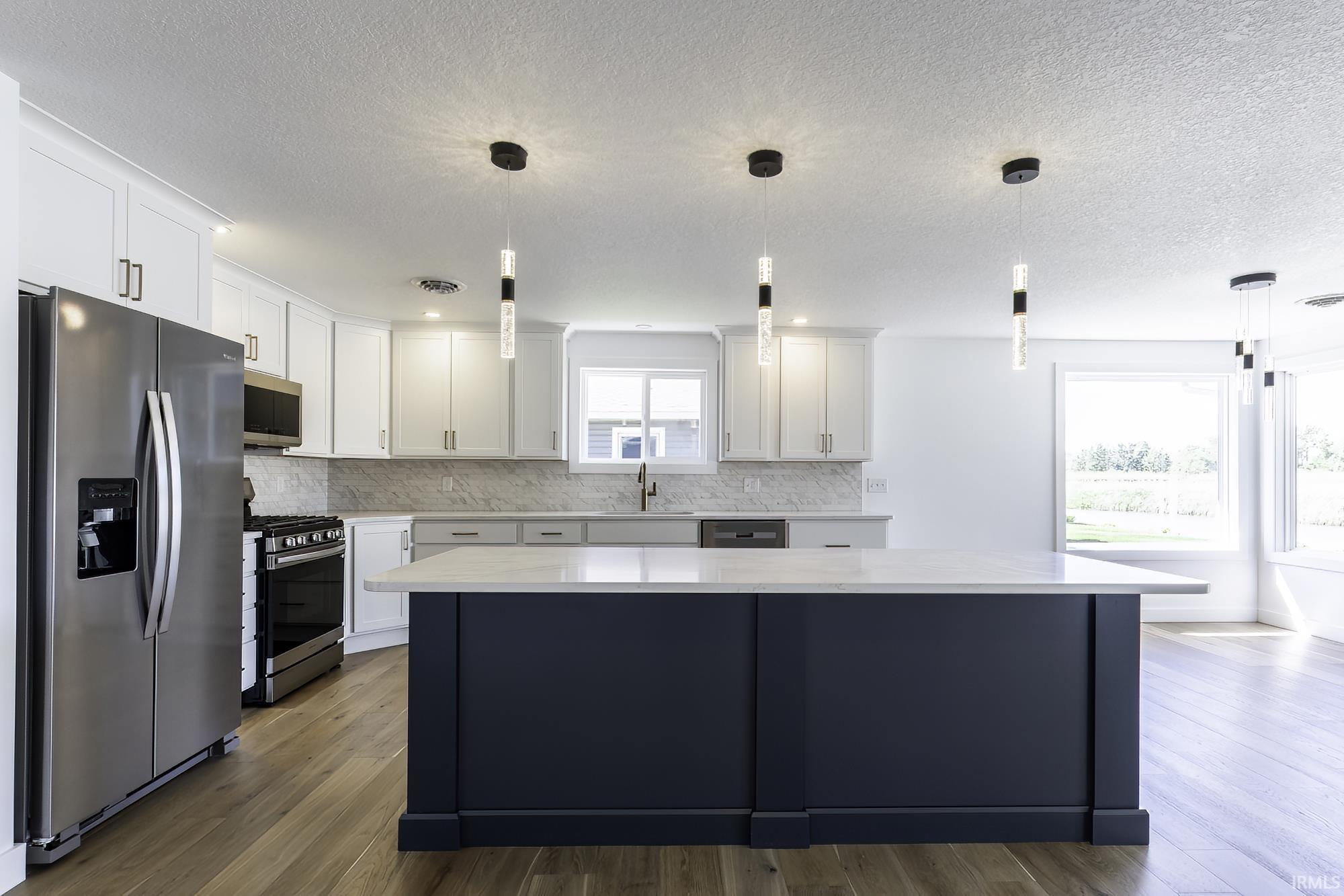 Image 3: Open-concept kitchen featuring a spacious central island with a light-toned countertop, white cabinetry with contemporary hardware, stainless steel appliances, modern pendant lighting, and wood-finish flooring, Kitchen