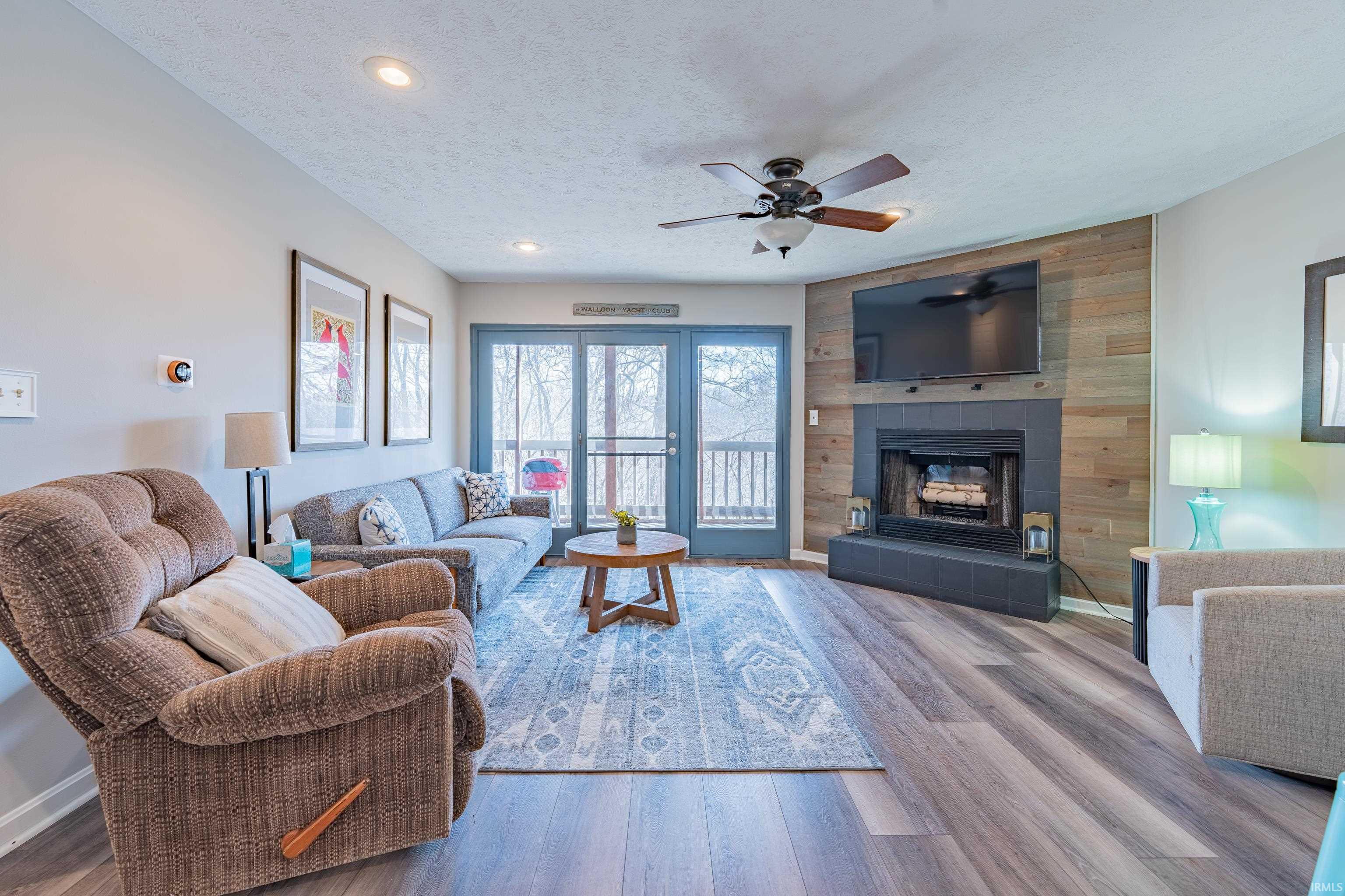 Image 1: Living room featuring wood finished floors, a fireplace, a textured ceiling, wooden walls, and ceiling fan, Living Room Image 1: Living room featuring wood finished floors, a fireplace, a textured ceiling, wooden walls, and ceiling fan, Living Room