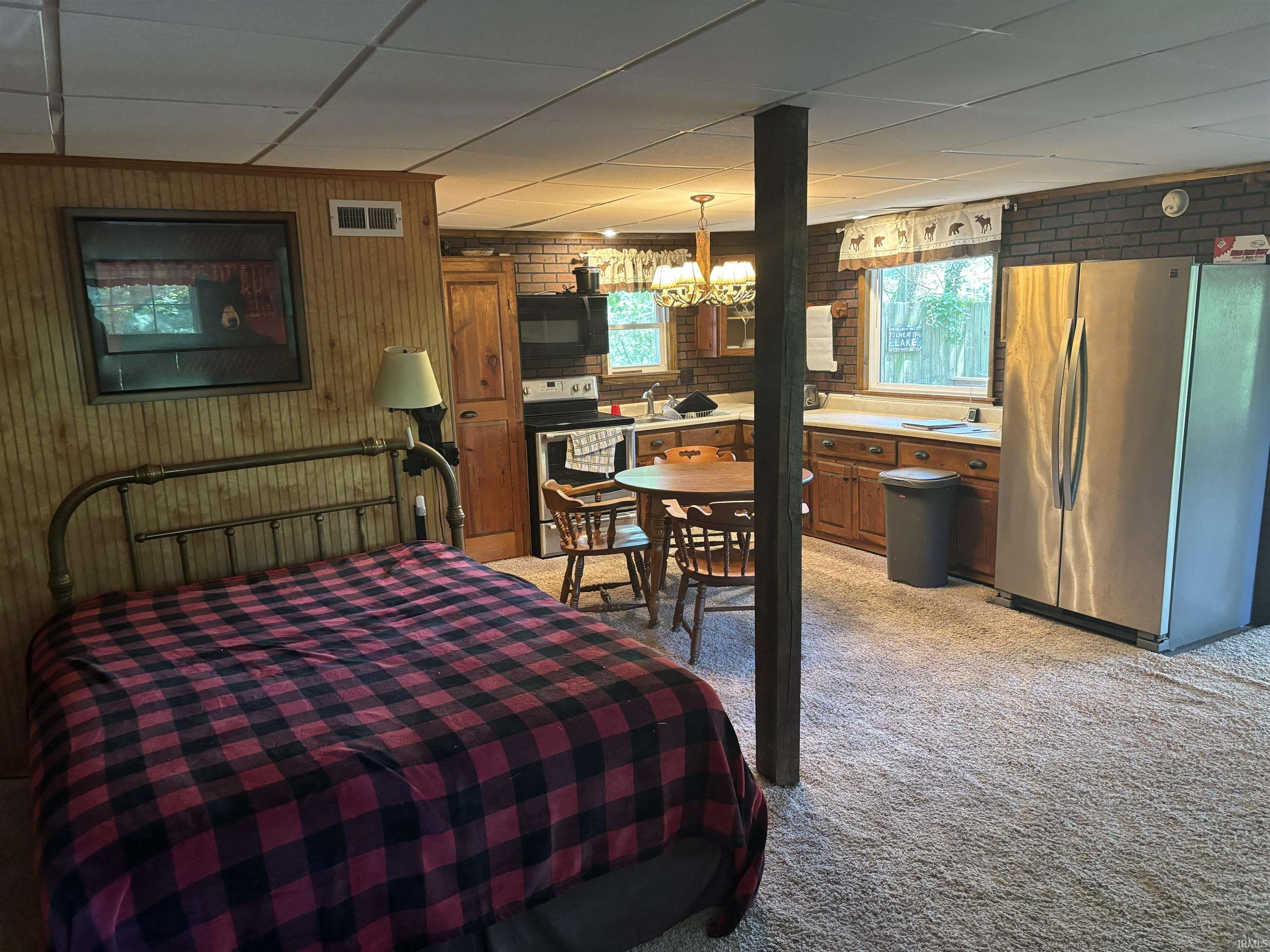 Image 3: Open-concept living space featuring wood-paneled walls, a compact kitchen, stainless steel refrigerator, and a window with outdoor scenery, Bedroom