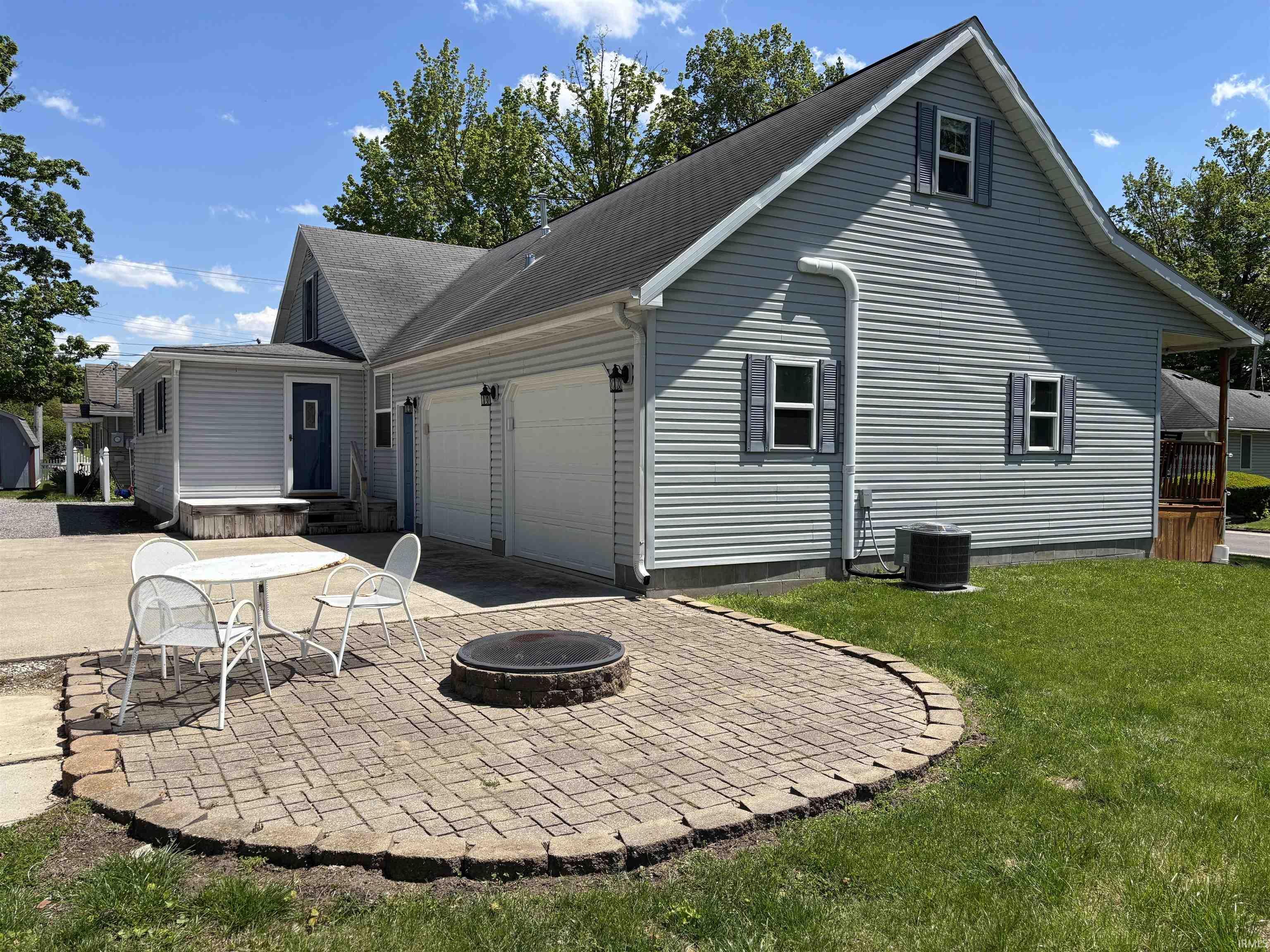 Image 2: Exterior featuring light blue siding, a two-car garage with white doors, and a paver patio with a built-in fire pit, Back Of Structure Image 2: Exterior featuring light blue siding, a two-car garage with white doors, and a paver patio with a built-in fire pit, Back Of Structure