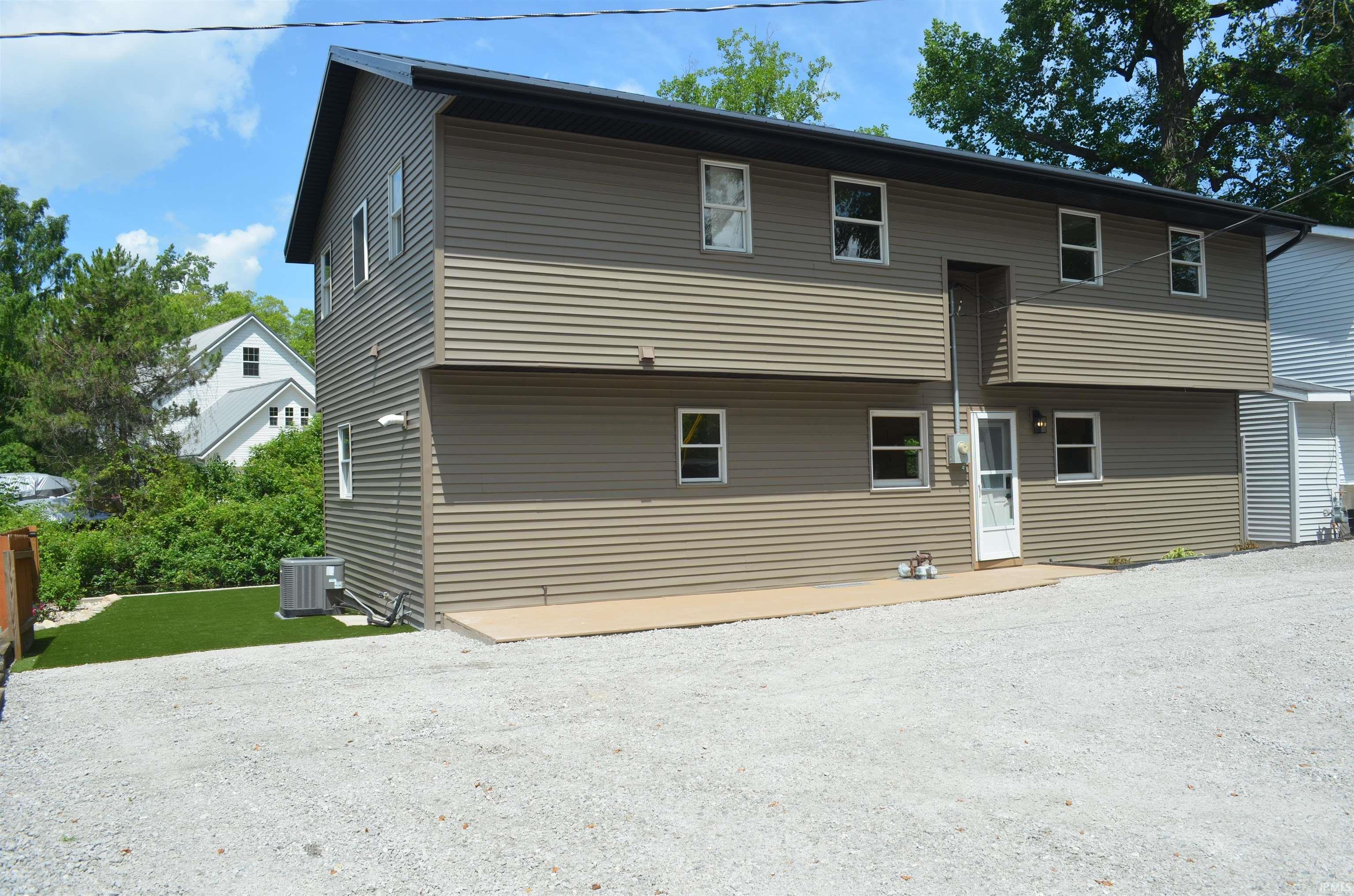 Image 1: Two-story residential structure featuring horizontal siding, multiple windows, a rear entry door, a gravel parking area, and an artificial turf section, Front Of Structure Image 1: Two-story residential structure featuring horizontal siding, multiple windows, a rear entry door, a gravel parking area, and an artificial turf section, Front Of Structure