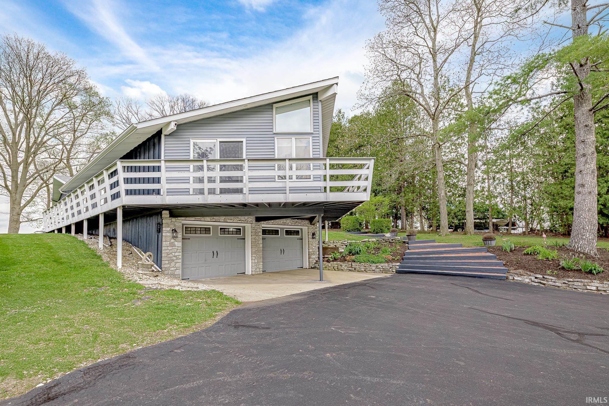 Image 3: Contemporary architectural style residence featuring a two-car garage with stone accents, an extended upper-level deck, a sloped asphalt driveway, and a terraced landscaped front yard with stone retaining walls, Front Of Structure