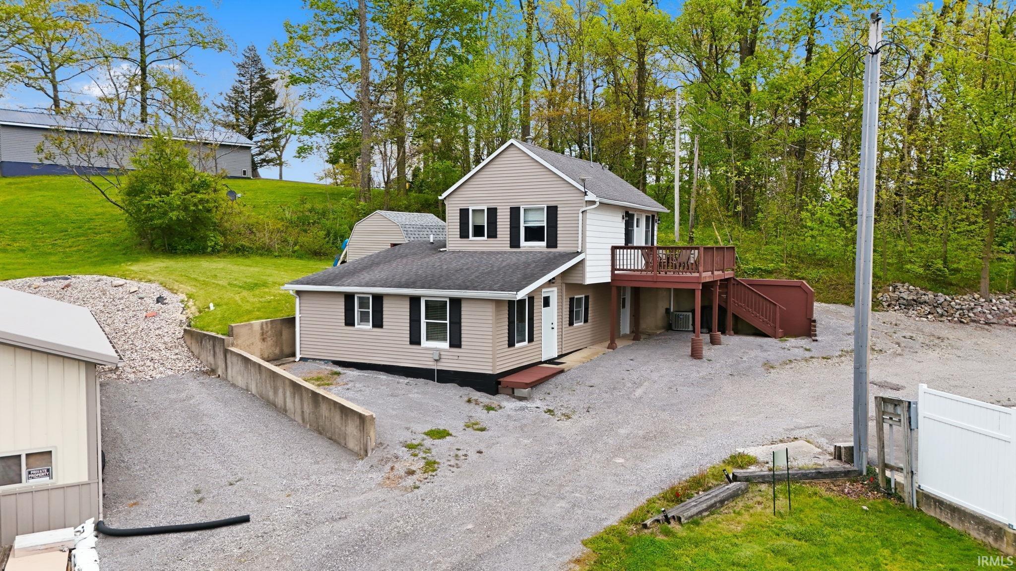 Image 3: Multi-level residence featuring tan siding with black shutters, a spacious gravel driveway, and a raised wooden deck with stairs, Front Of Structure Image 3: Multi-level residence featuring tan siding with black shutters, a spacious gravel driveway, and a raised wooden deck with stairs, Front Of Structure