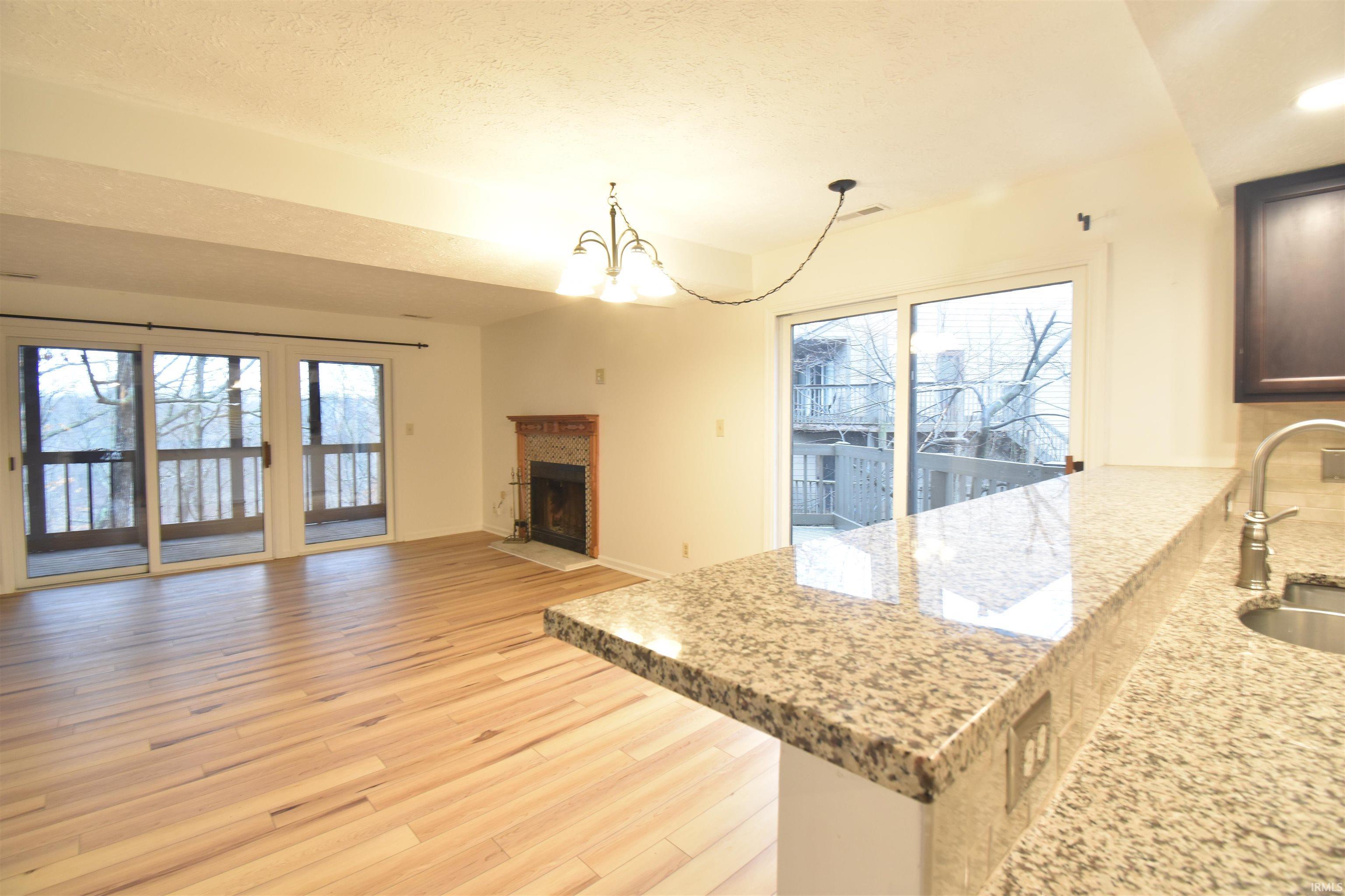 Image 1: Kitchen featuring light stone countertops, hanging lights, a fireplace with flush hearth, light wood-style floors, and plenty of natural light, Great Room Image 1: Kitchen featuring light stone countertops, hanging lights, a fireplace with flush hearth, light wood-style floors, and plenty of natural light, Great Room