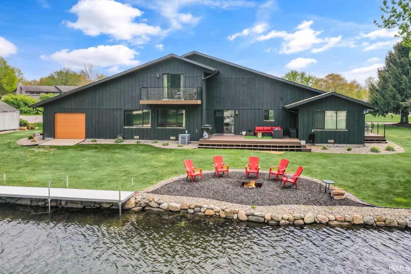 Image 3: Waterfront property featuring a dark-toned exterior with a wood deck, a private dock extending into the water, a waterside fire pit area with stone edging, and a second-story balcony, Back Of Structure Image 3: Waterfront property featuring a dark-toned exterior with a wood deck, a private dock extending into the water, a waterside fire pit area with stone edging, and a second-story balcony, Back Of Structure