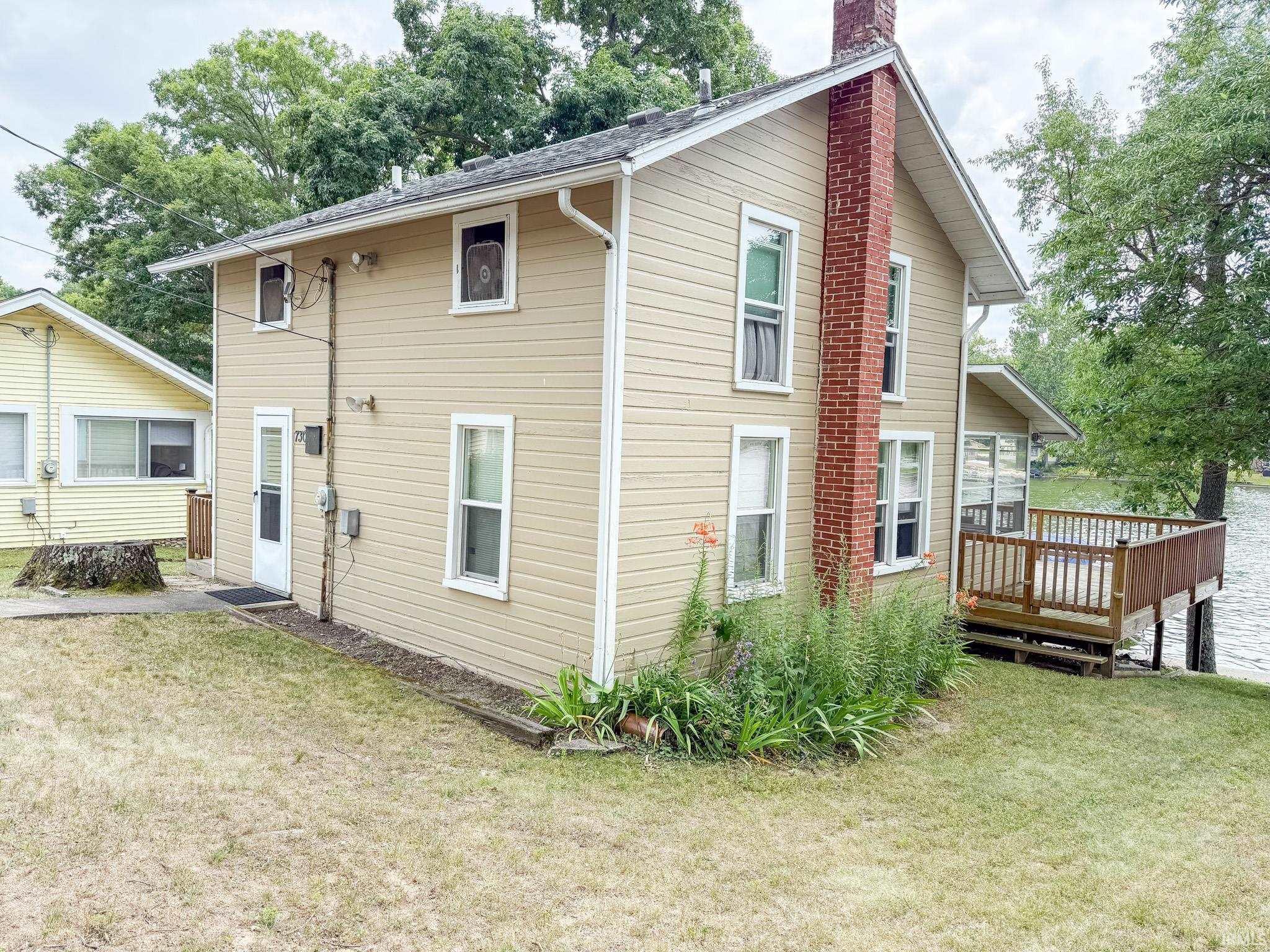 Image 2: Exterior featuring beige horizontal siding, a red brick chimney, white trim, a brown wood deck over water, and mature trees in the surrounding landscape, Back Of Structure Image 2: Exterior featuring beige horizontal siding, a red brick chimney, white trim, a brown wood deck over water, and mature trees in the surrounding landscape, Back Of Structure