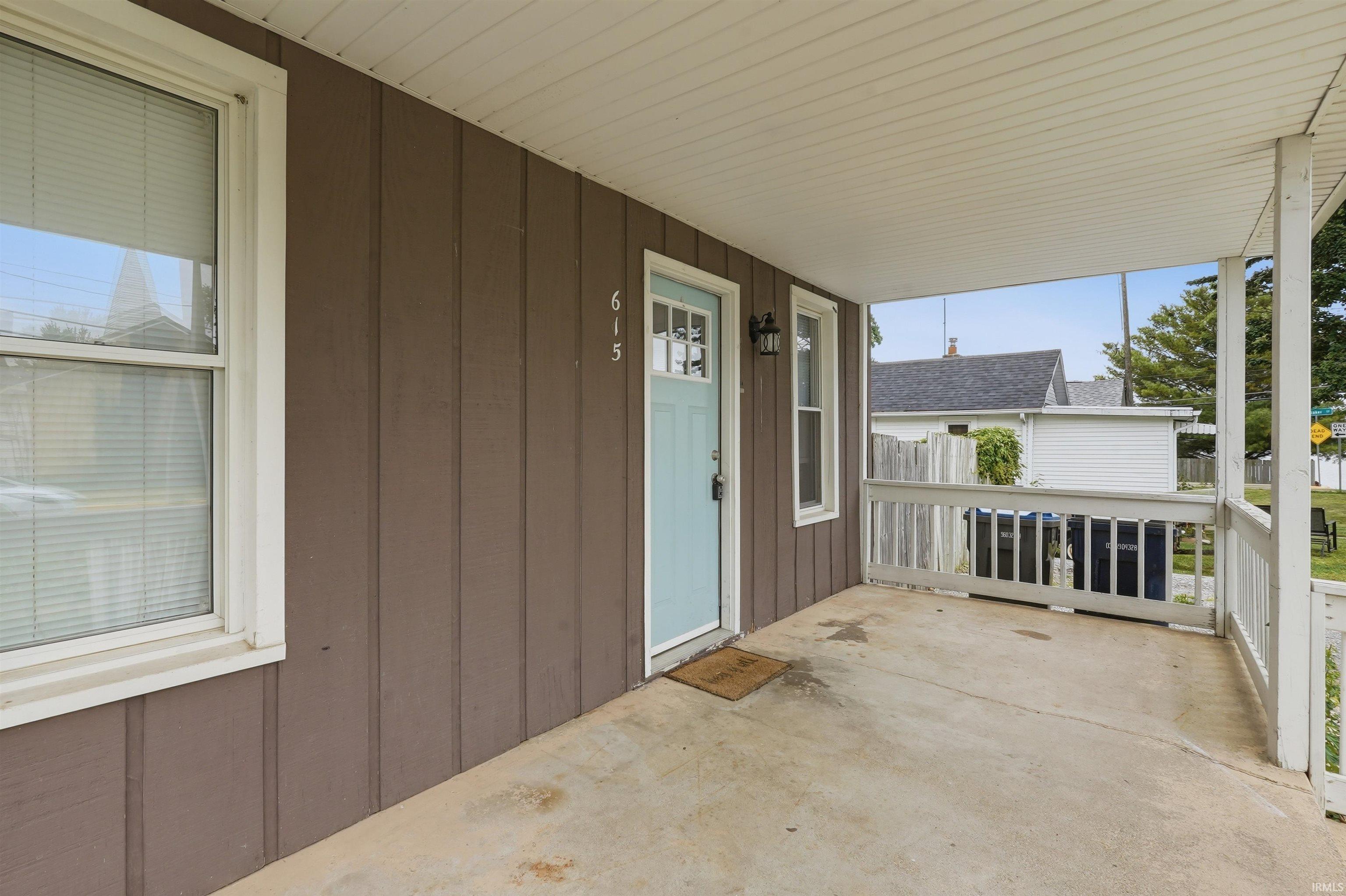Image 3: Covered front porch featuring a light blue entry door with decorative glass, concrete flooring, vertical brown siding, and white railing, Patio