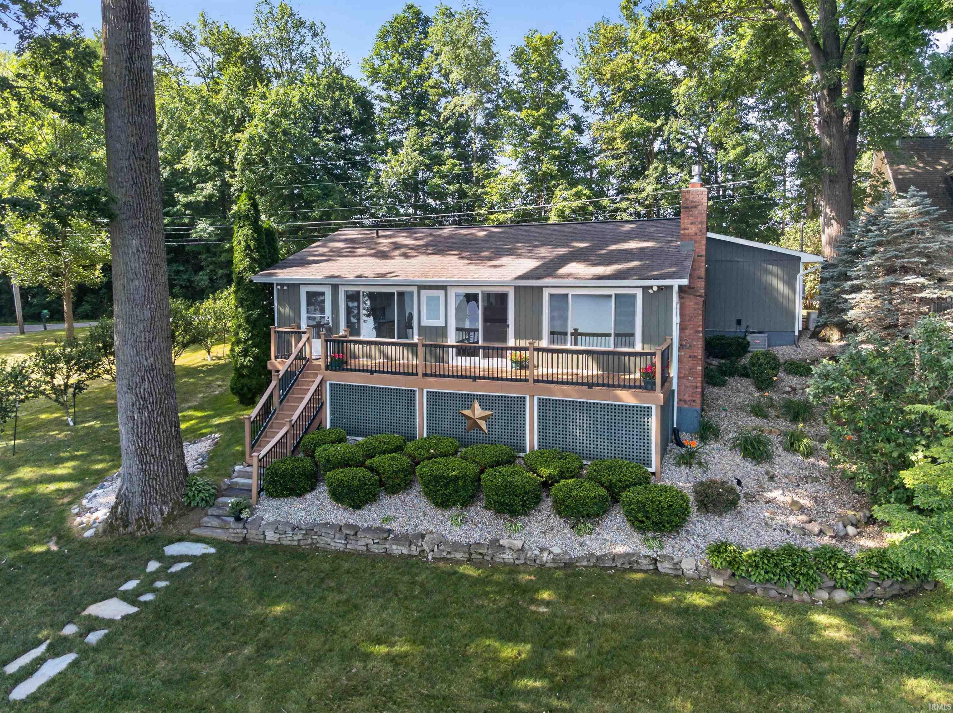Image 1: Waterfront property featuring a spacious deck with dark railings, a prominent brick chimney, and a landscaped yard with mature trees, back Image 1: Waterfront property featuring a spacious deck with dark railings, a prominent brick chimney, and a landscaped yard with mature trees, back