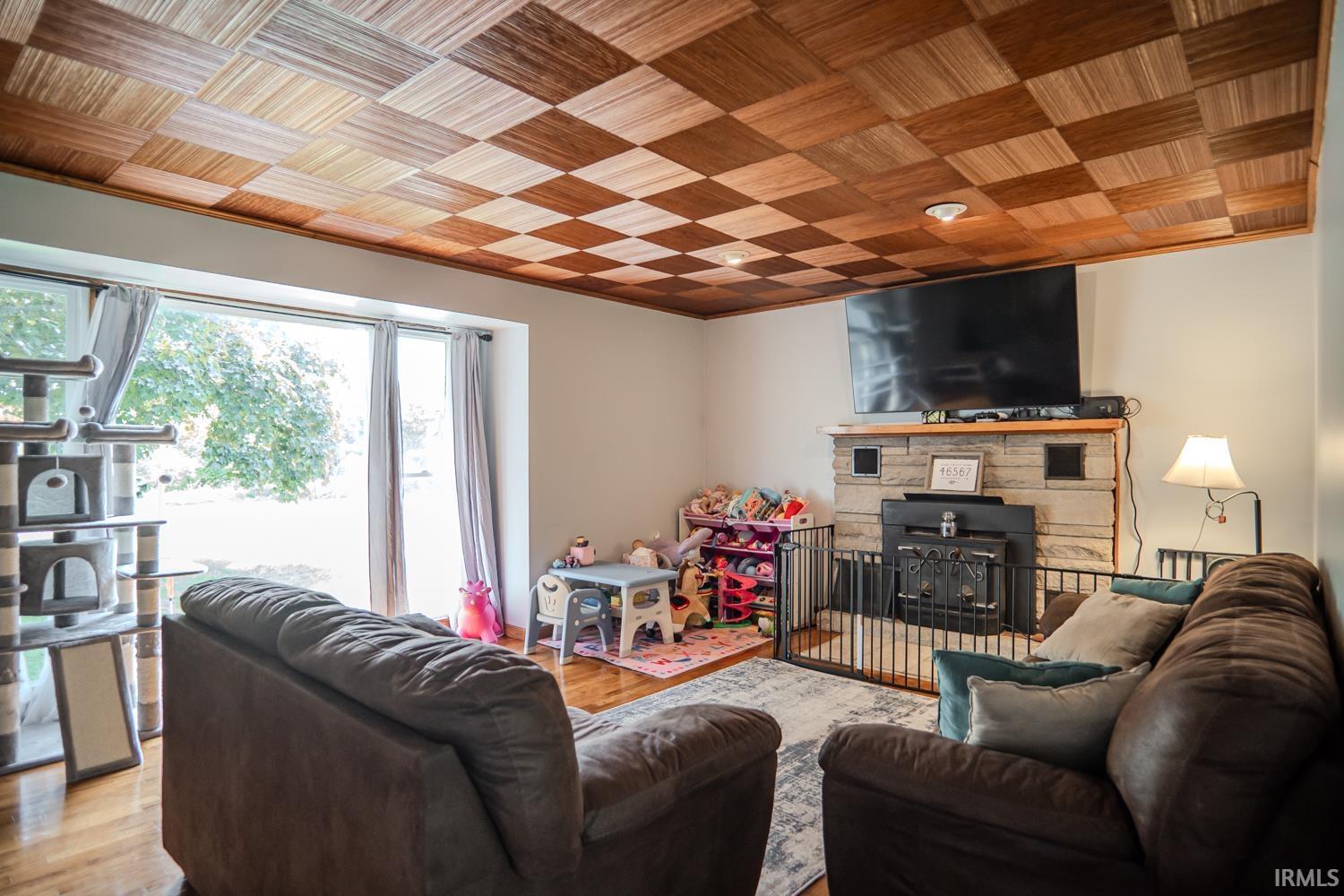 Image 2: Living room featuring a checkerboard wood ceiling, stone-surround fireplace with a hearth, and large picture windows, Living Room Image 2: Living room featuring a checkerboard wood ceiling, stone-surround fireplace with a hearth, and large picture windows, Living Room
