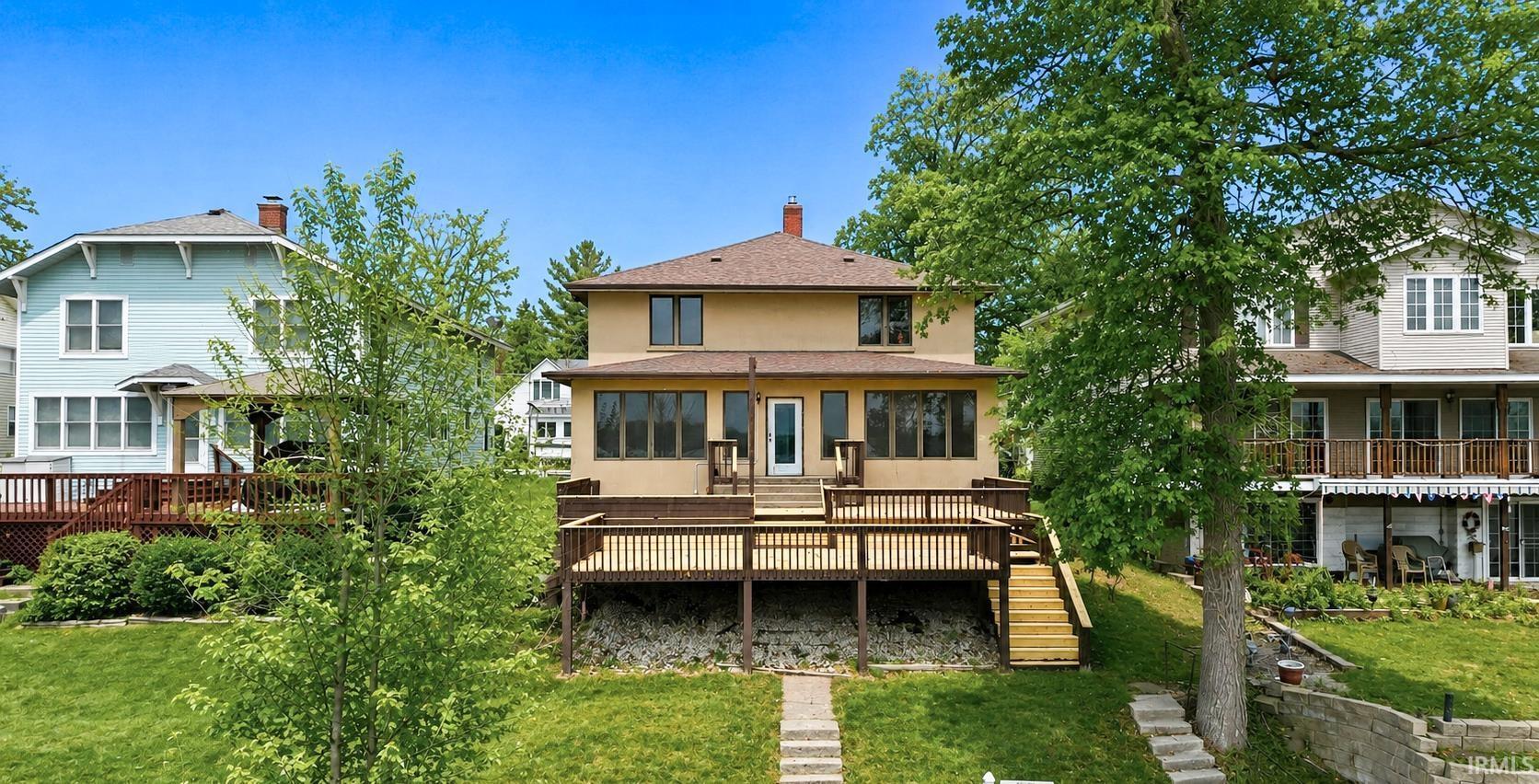 Image 1: Two-story stucco property featuring a multi-level wood deck with wood railings, a sunroom extension, and multiple windows across the rear facade, Back Of Structure Image 1: Two-story stucco property featuring a multi-level wood deck with wood railings, a sunroom extension, and multiple windows across the rear facade, Back Of Structure