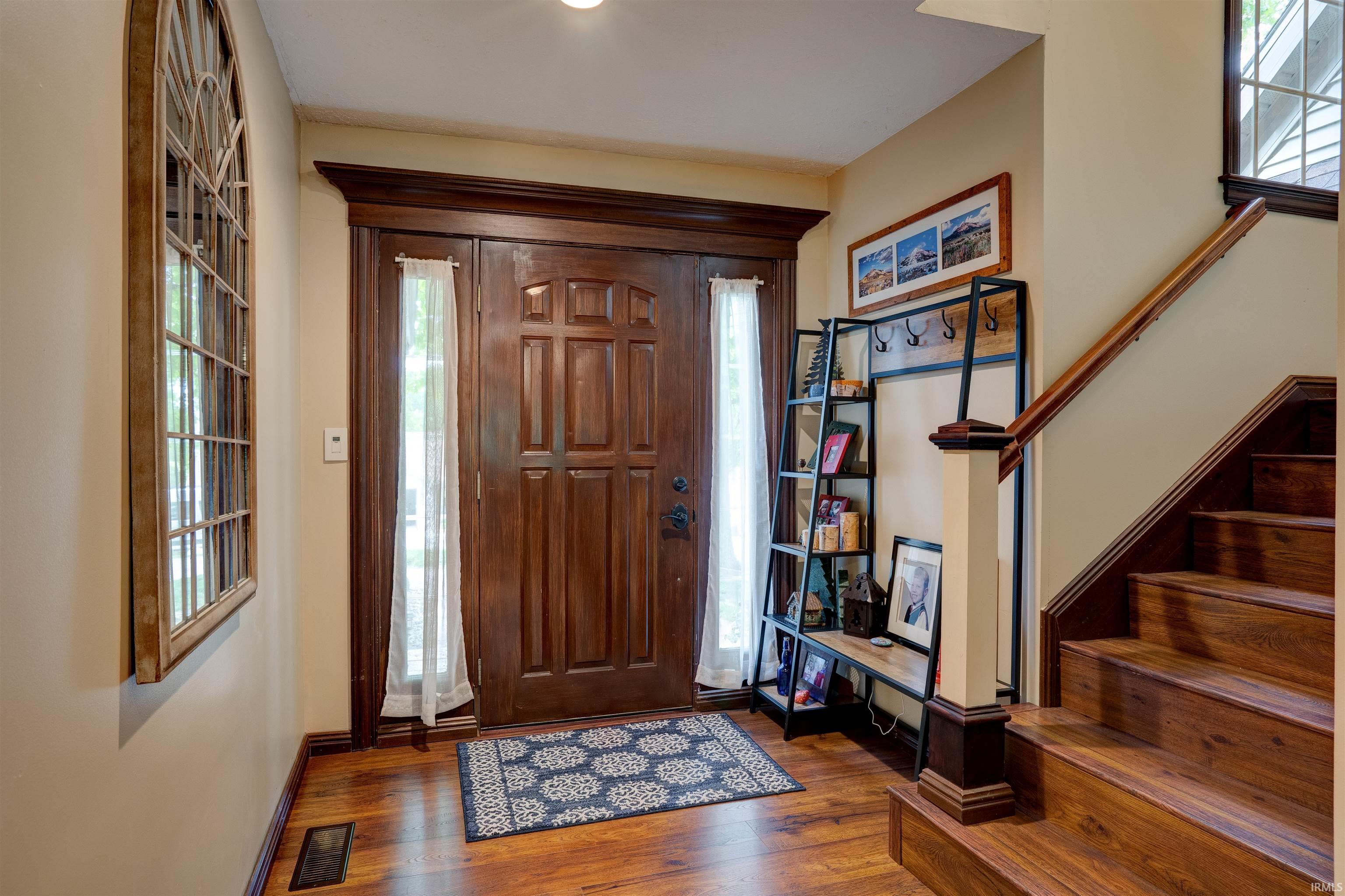 Image 2: Entryway with rich wood-finish flooring, a paneled wood entry door, and a matching wood staircase with a substantial newel post, Entrance Foyer Image 2: Entryway with rich wood-finish flooring, a paneled wood entry door, and a matching wood staircase with a substantial newel post, Entrance Foyer