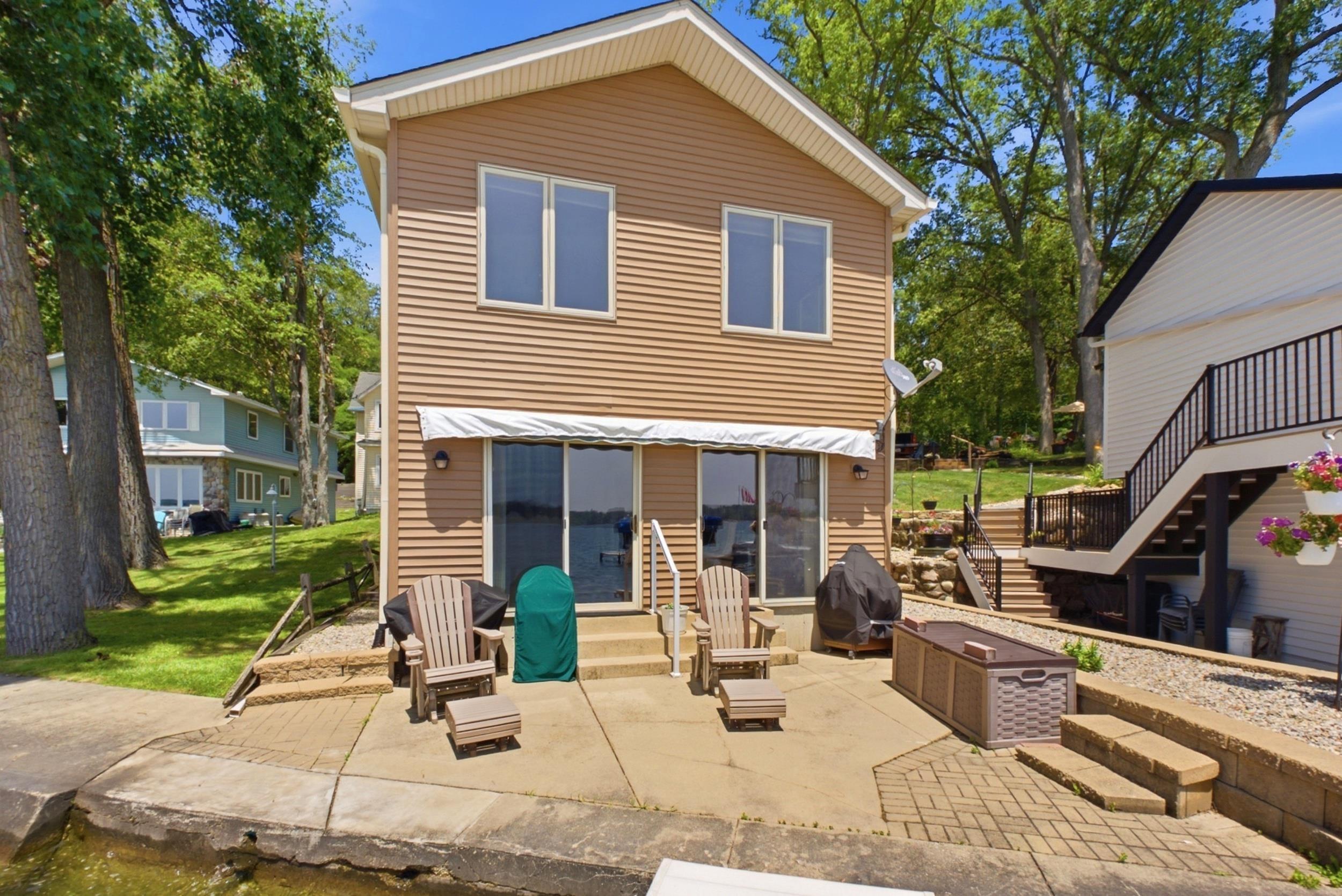 Image 1: Waterfront property featuring a two-story residence with tan horizontal siding, white trim, and a white awning above a lower-level patio, Back Of Structure Image 1: Waterfront property featuring a two-story residence with tan horizontal siding, white trim, and a white awning above a lower-level patio, Back Of Structure