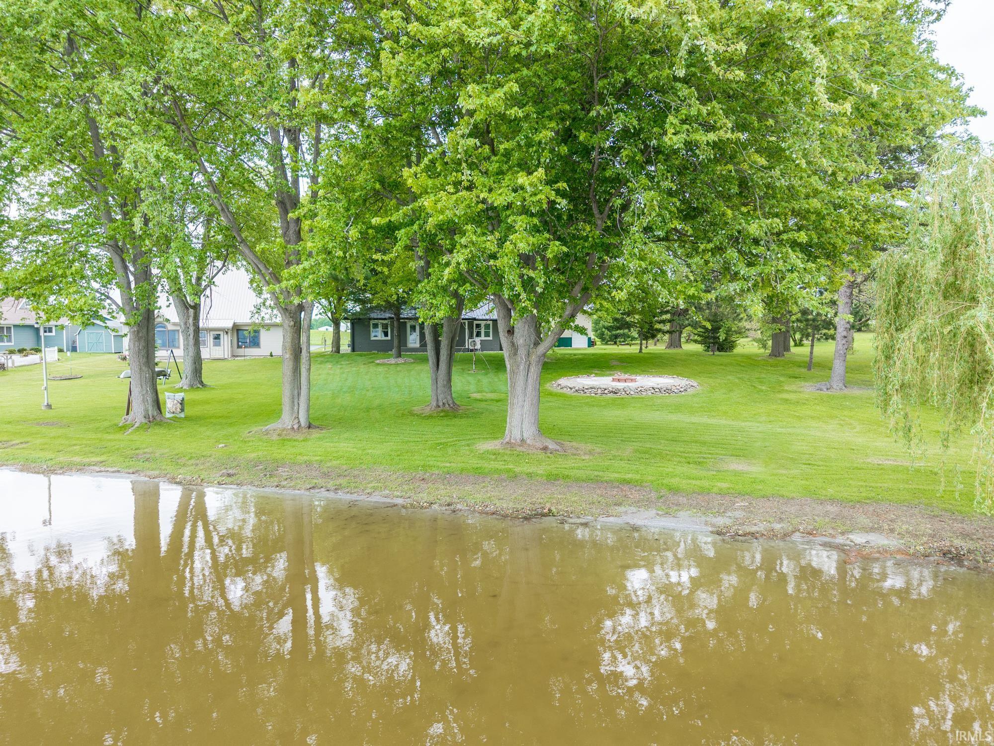 Image 3: Expansive green lawn featuring mature trees and a circular stone fire pit, with water frontage, 150 ft Lakefront home on Fish Lake .68 acre lot renovated turn key home and 30x40 pole building