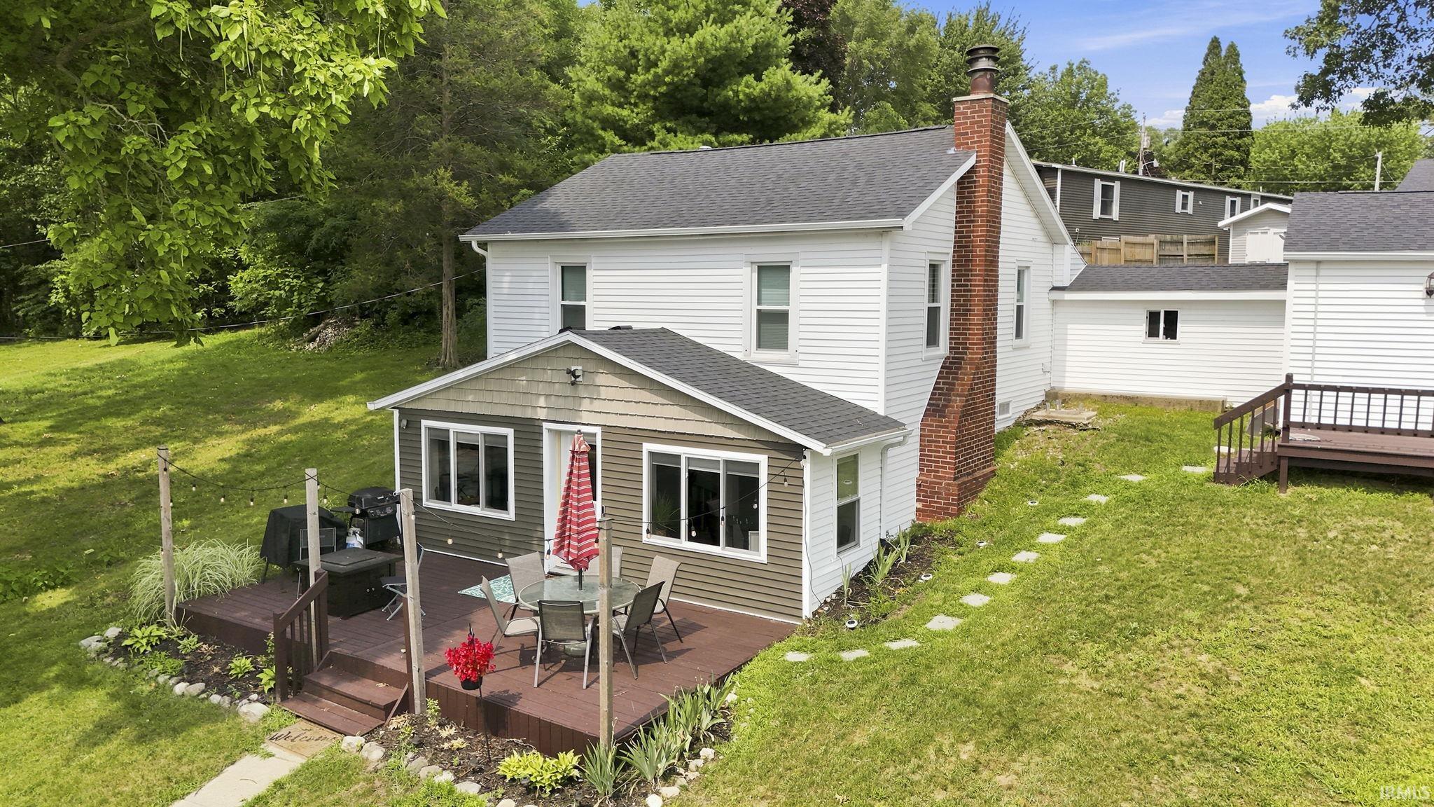 Image 0: White siding residence featuring a large rear deck, a brick chimney, a detached garage with white siding, and a sloped grass yard, Front Of Structure