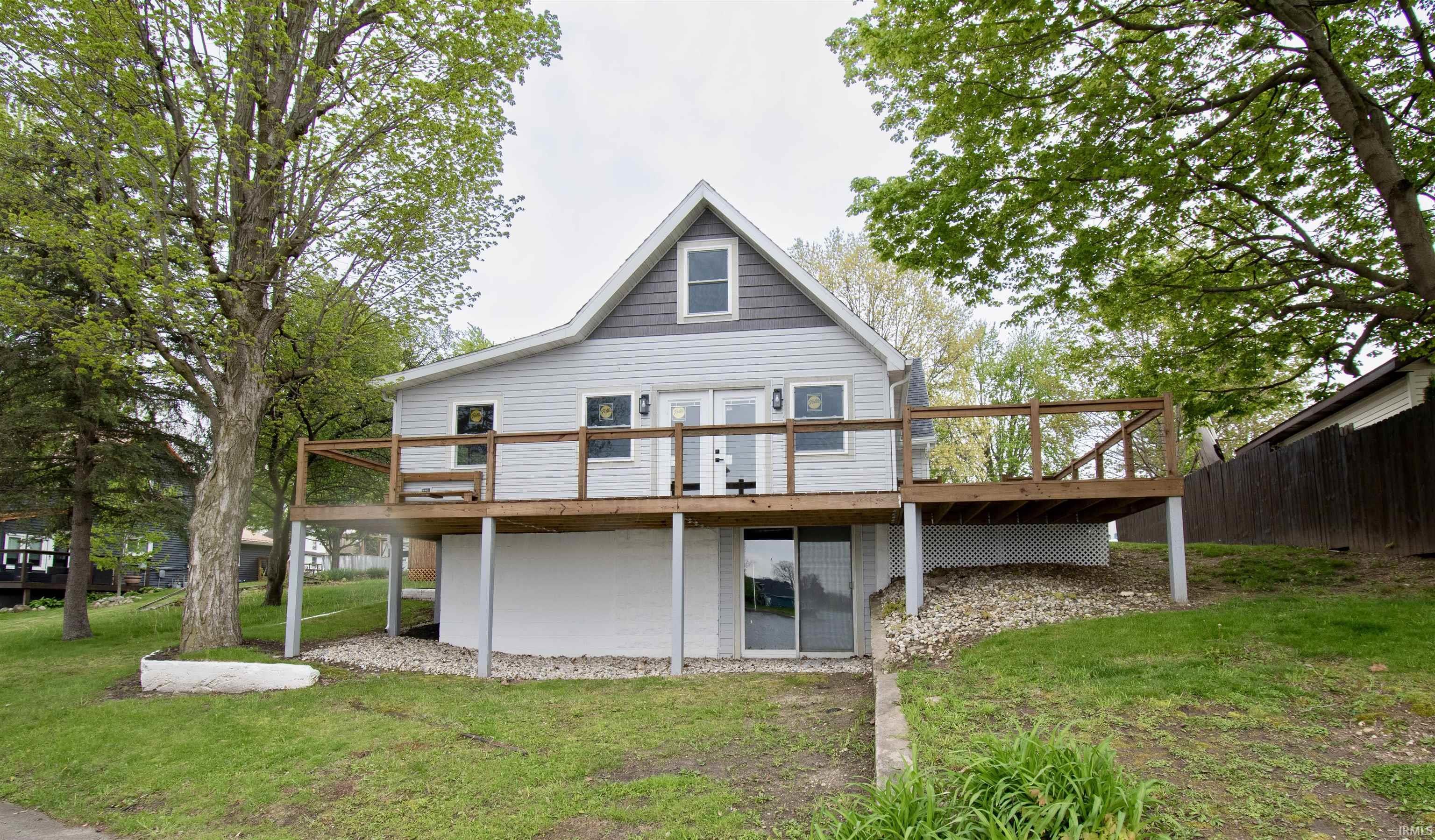 Image 0: Two-story residence featuring light gray siding, an expansive elevated wood deck, a prominent white foundation, a lower-level sliding glass door, and mature trees providing natural landscaping, Back Of Structure Image 0: Two-story residence featuring light gray siding, an expansive elevated wood deck, a prominent white foundation, a lower-level sliding glass door, and mature trees providing natural landscaping, Back Of Structure