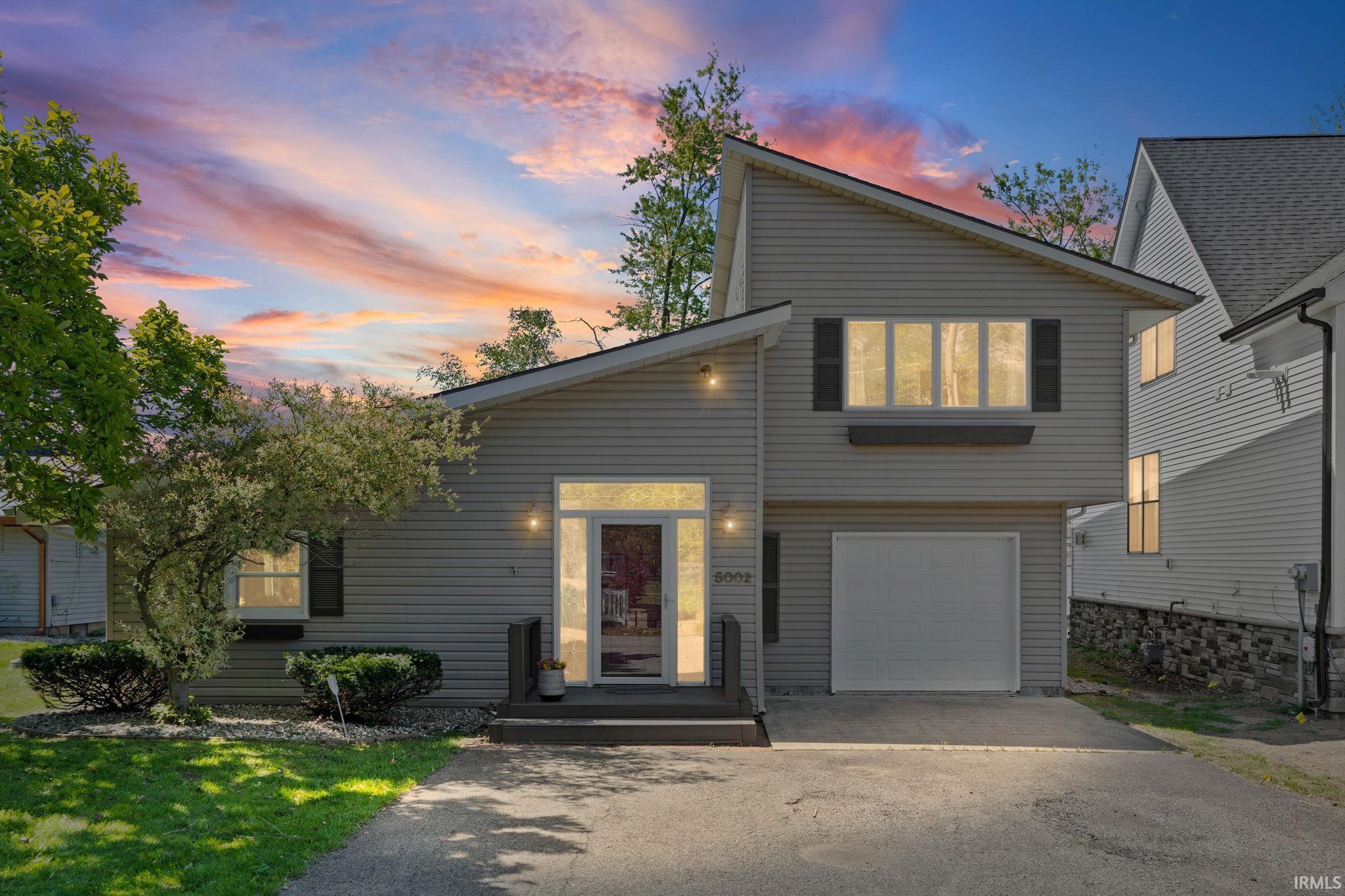 Image 1: Exterior featuring light gray siding, an attached garage, a prominent front door with sidelights, a landscaped front yard with a mature tree, and a paved driveway, Front Of Structure