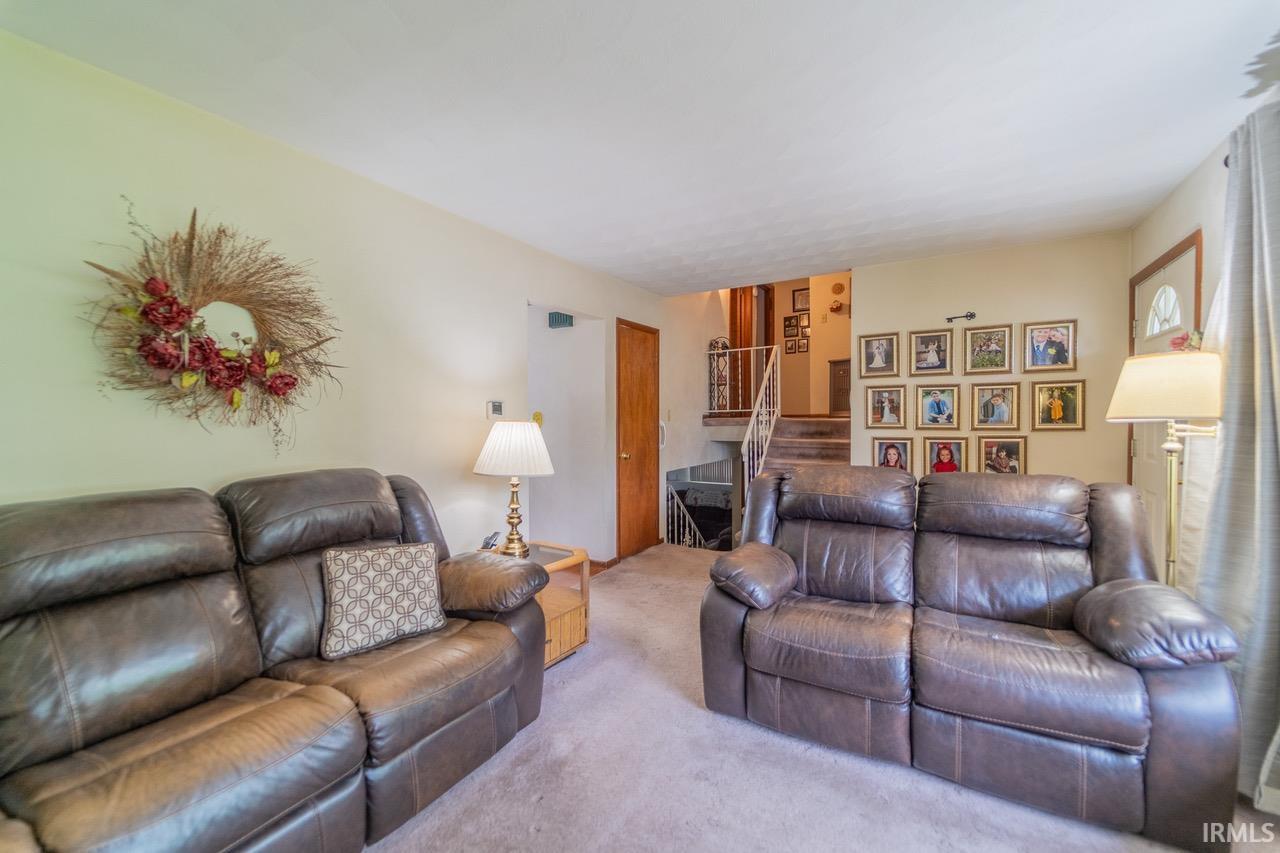 Image 2: Carpeted living area featuring a wood-finish door, staircase with white railings, and light-toned walls, Living Room Image 2: Carpeted living area featuring a wood-finish door, staircase with white railings, and light-toned walls, Living Room