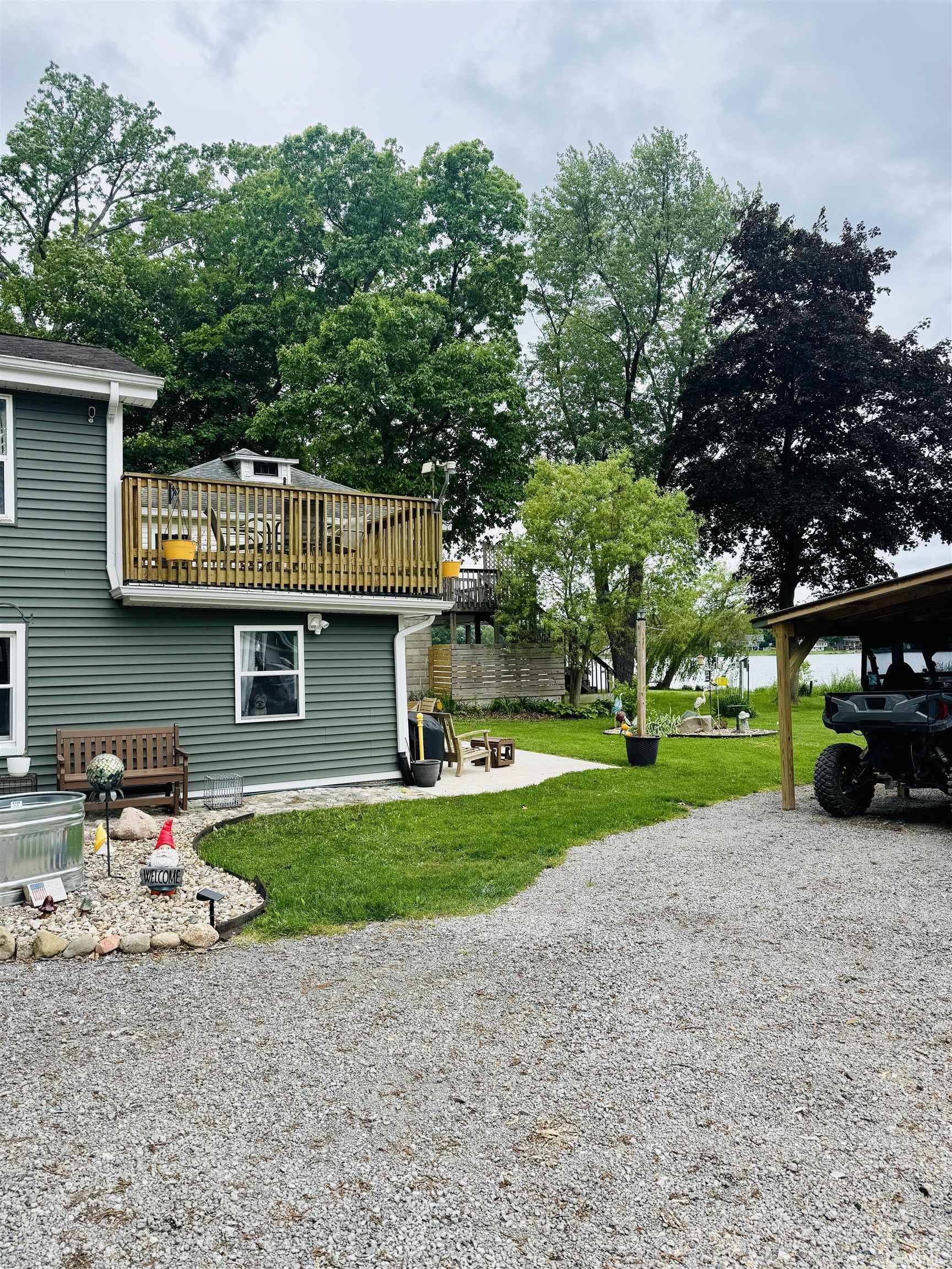 Image 2: Exterior featuring a second-story wood deck, green siding, a gravel driveway, a grassy lawn, and mature trees, Yard Image 2: Exterior featuring a second-story wood deck, green siding, a gravel driveway, a grassy lawn, and mature trees, Yard