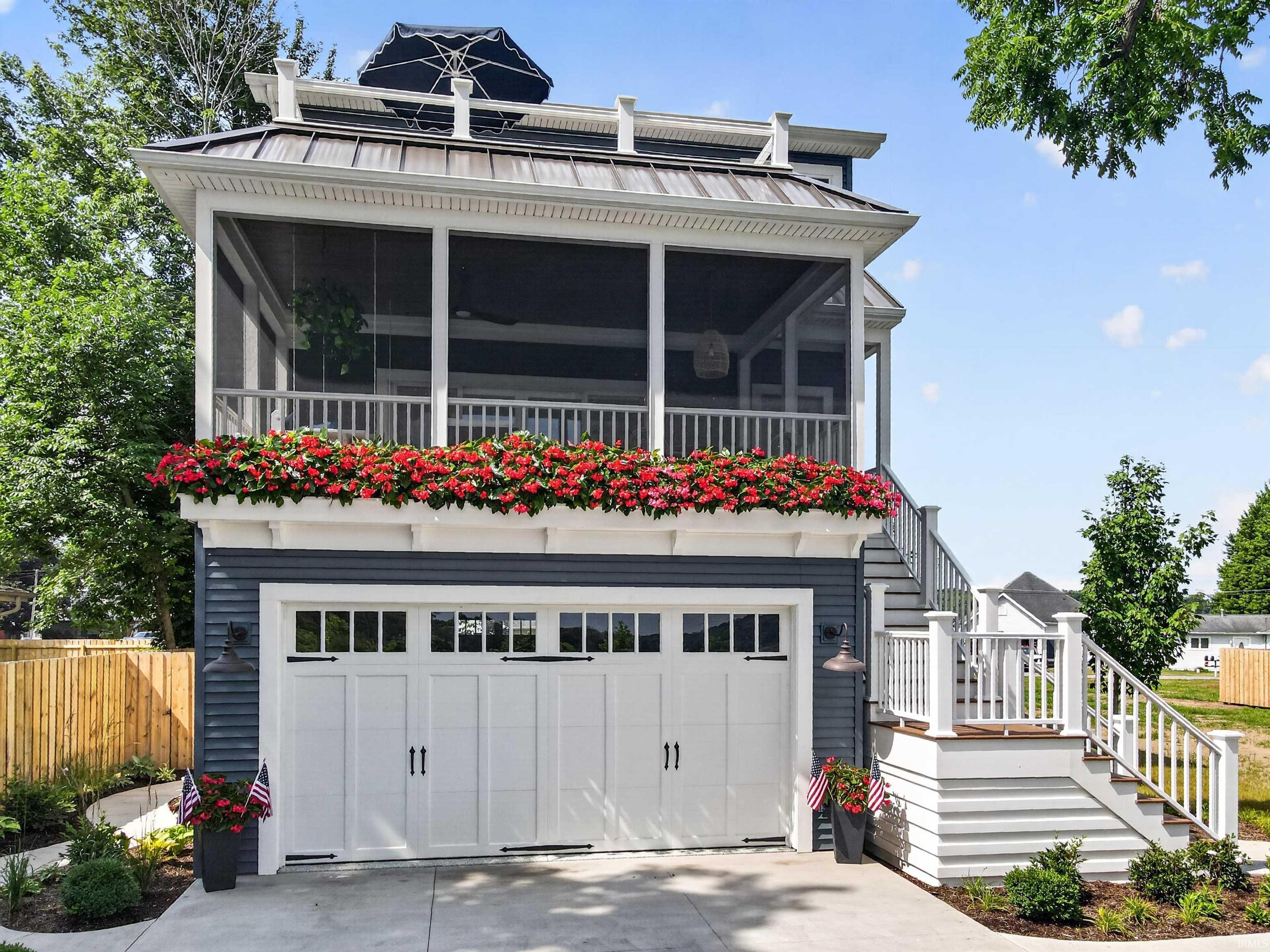 Image 0: Two-car garage with white paneled doors, screened porch with white railing, exterior staircase with white railings, dark blue siding, and a metal roof, Front Of Structure Image 0: Two-car garage with white paneled doors, screened porch with white railing, exterior staircase with white railings, dark blue siding, and a metal roof, Front Of Structure
