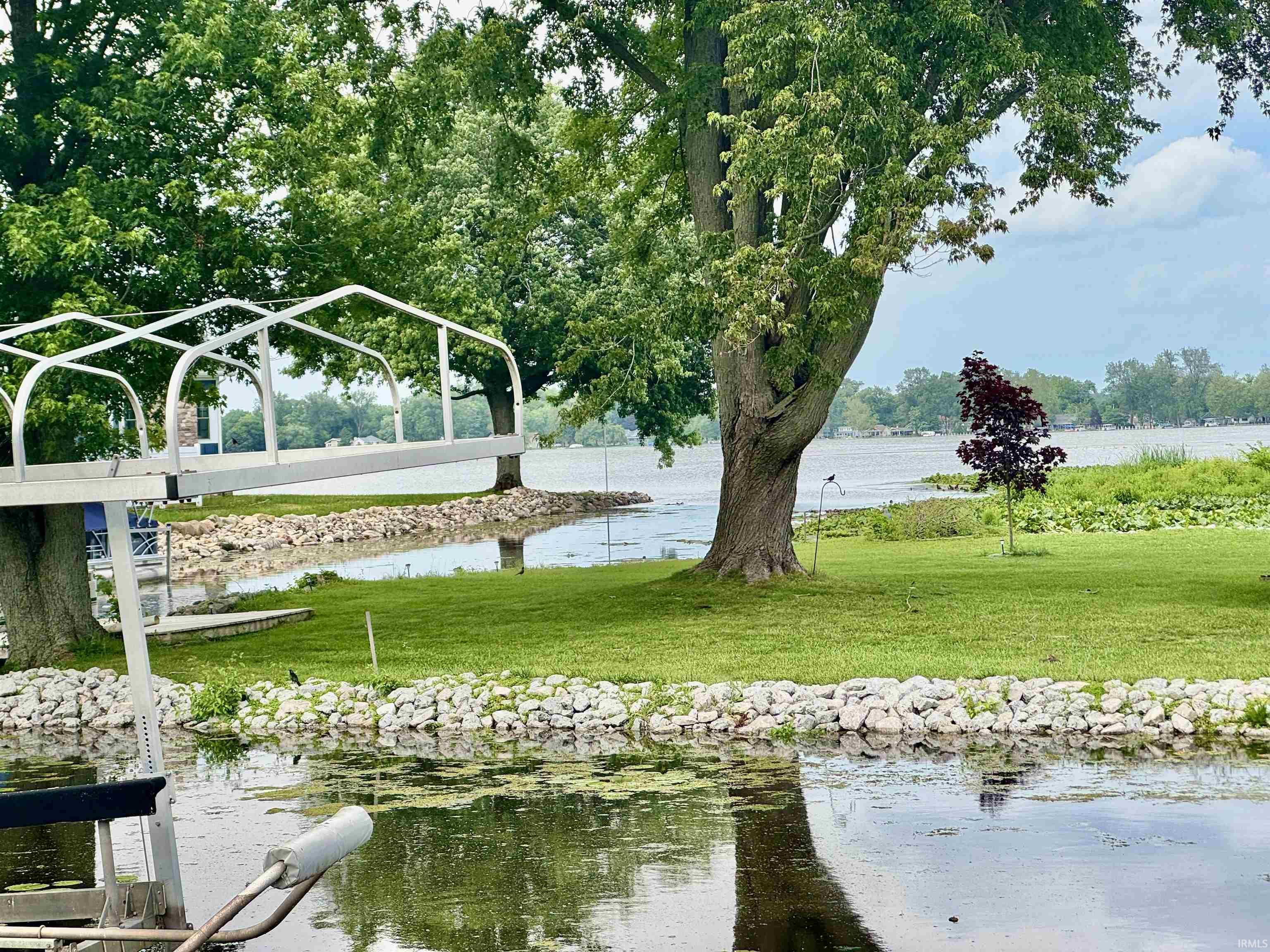 Image 2: Waterfront property featuring a boat hoist, lush green lawn, mature trees, and a protective rock shoreline, Yard