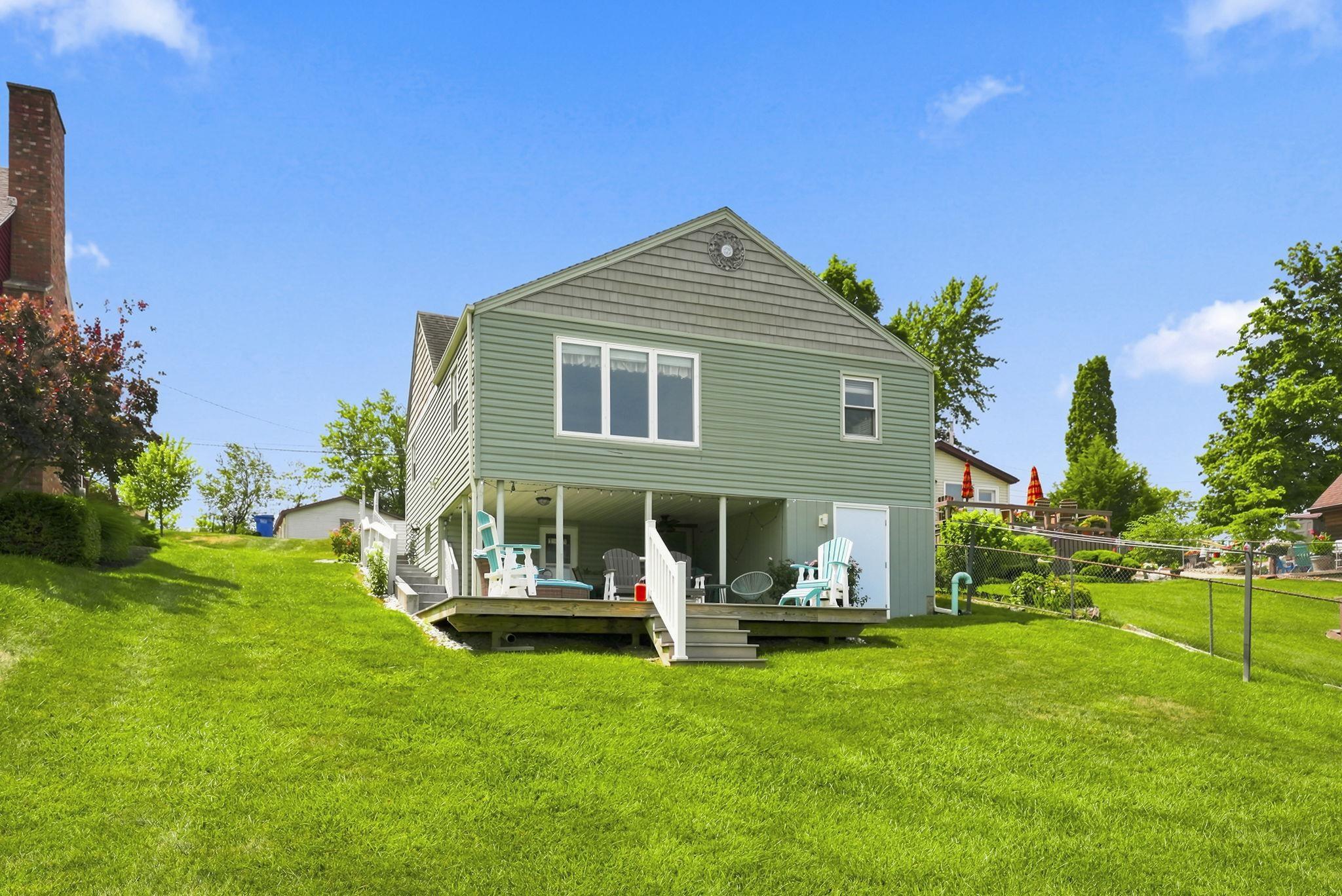 Image 1: Rear exterior featuring green siding, a lower-level covered patio, new composite deck and expansive yard to waterfront, Lakefront Side of cottage Image 1: Rear exterior featuring green siding, a lower-level covered patio, new composite deck and expansive yard to waterfront, Lakefront Side of cottage