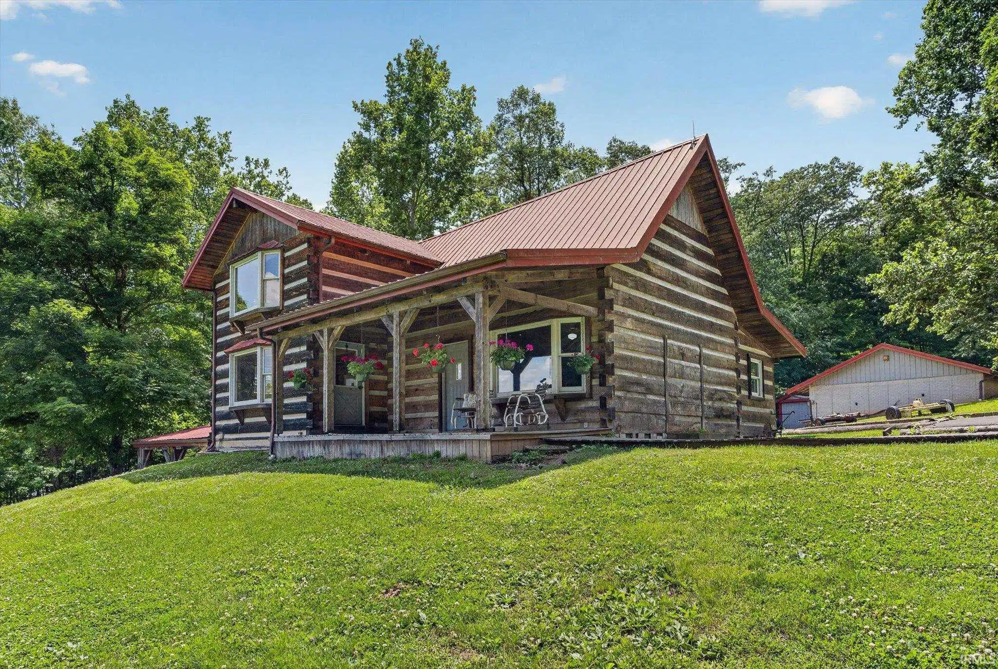 Image 0: Rustic log cabin residence featuring a metal roof, covered porch with natural wood posts, bay windows, and a detached outbuilding, Front Of Structure Image 0: Rustic log cabin residence featuring a metal roof, covered porch with natural wood posts, bay windows, and a detached outbuilding, Front Of Structure