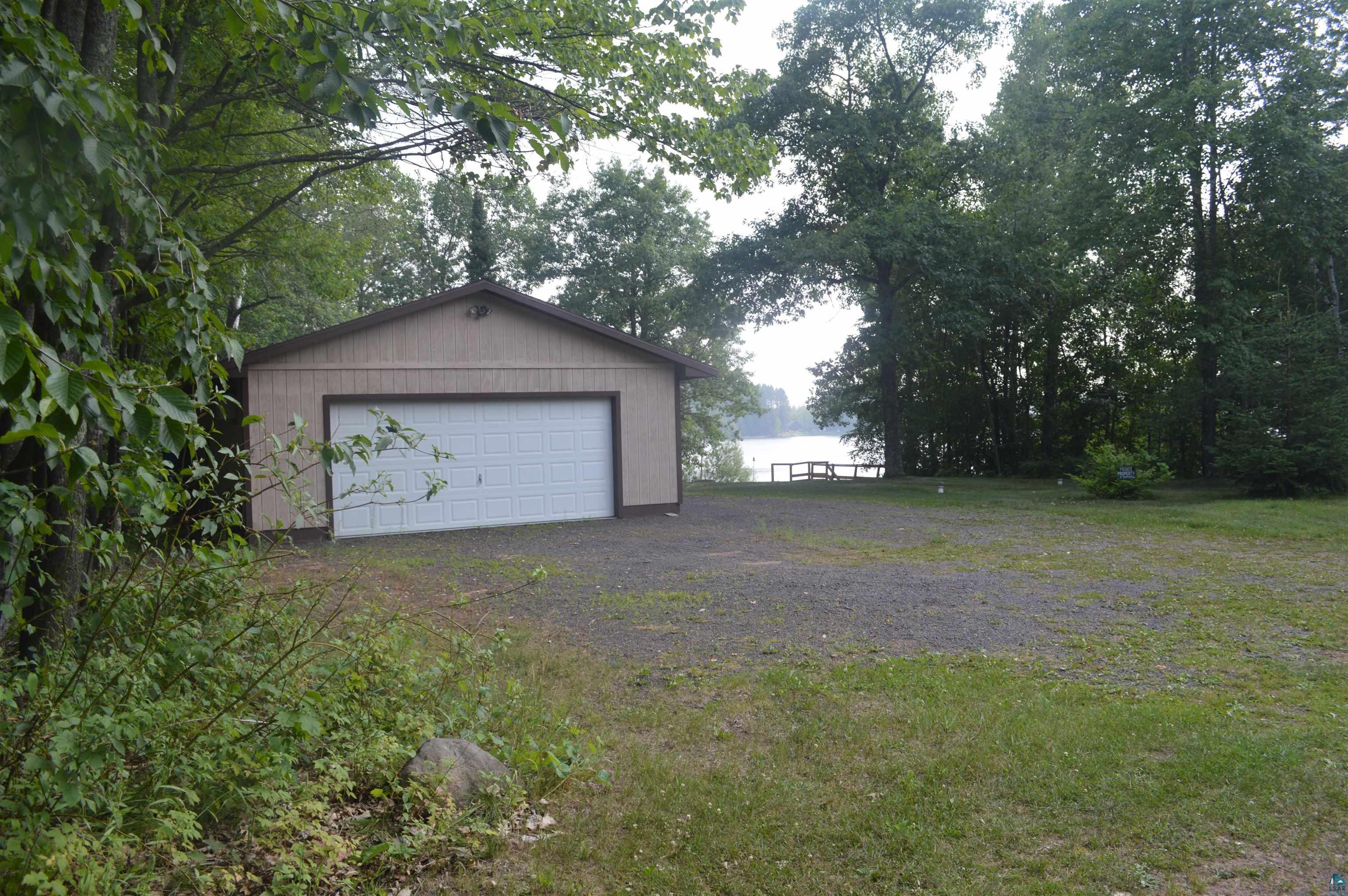 Image 1: Detached garage with a white overhead door, gravel driveway, and a lake visible through mature trees, Garage