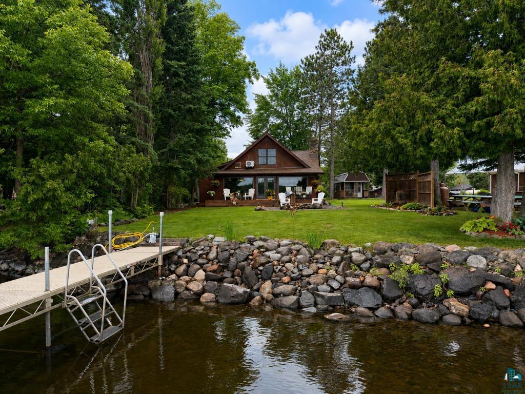 Image 2: Waterfront property featuring a private dock with ladder, a rock-lined shore, a spacious green lawn, a wood-sided residence with an expansive deck, and mature perimeter trees, Dock