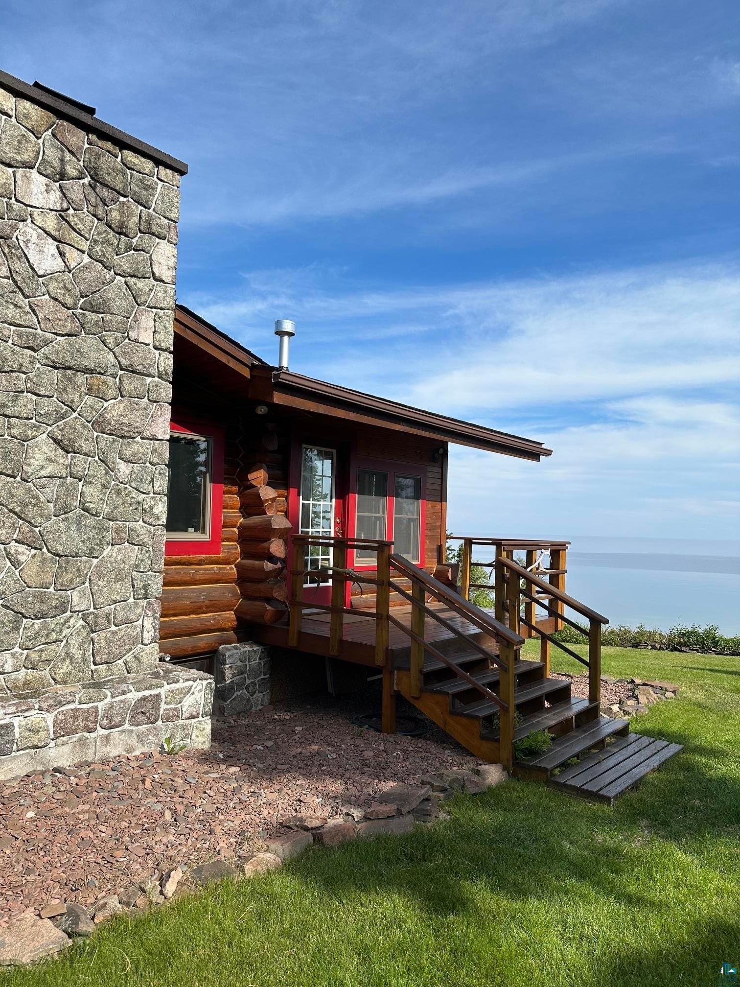 Image 2: Log cabin exterior featuring a multi-level wood deck, stone-finish chimney, red window frames, and a gravel walkway, Side Of Structure