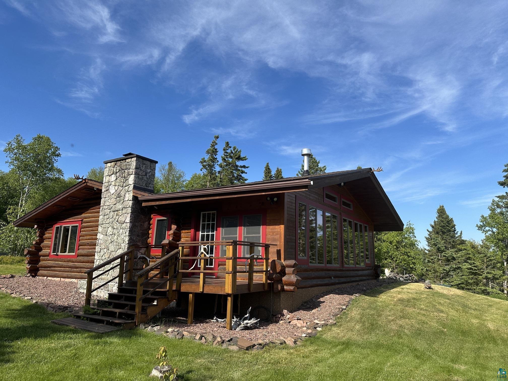 Image 3: Log cabin exterior featuring a stone chimney, extensive window line, wood deck with railing, and a sloping grassy yard, Back Of Structure