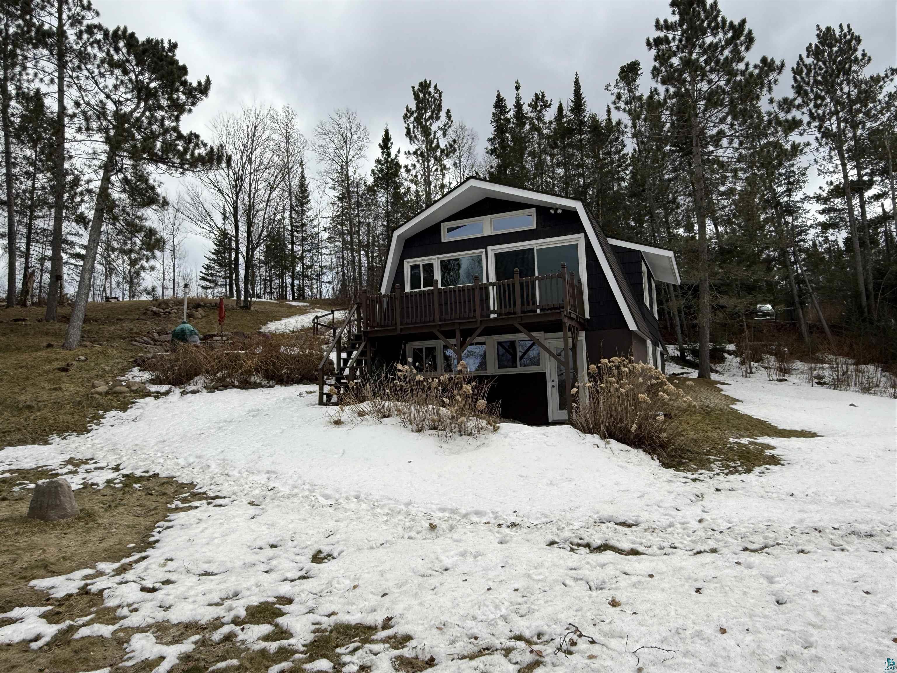 Image 1: Snow covered rear of property with a gambrel roof, a wooden deck, and view of scattered trees, Back Of Structure