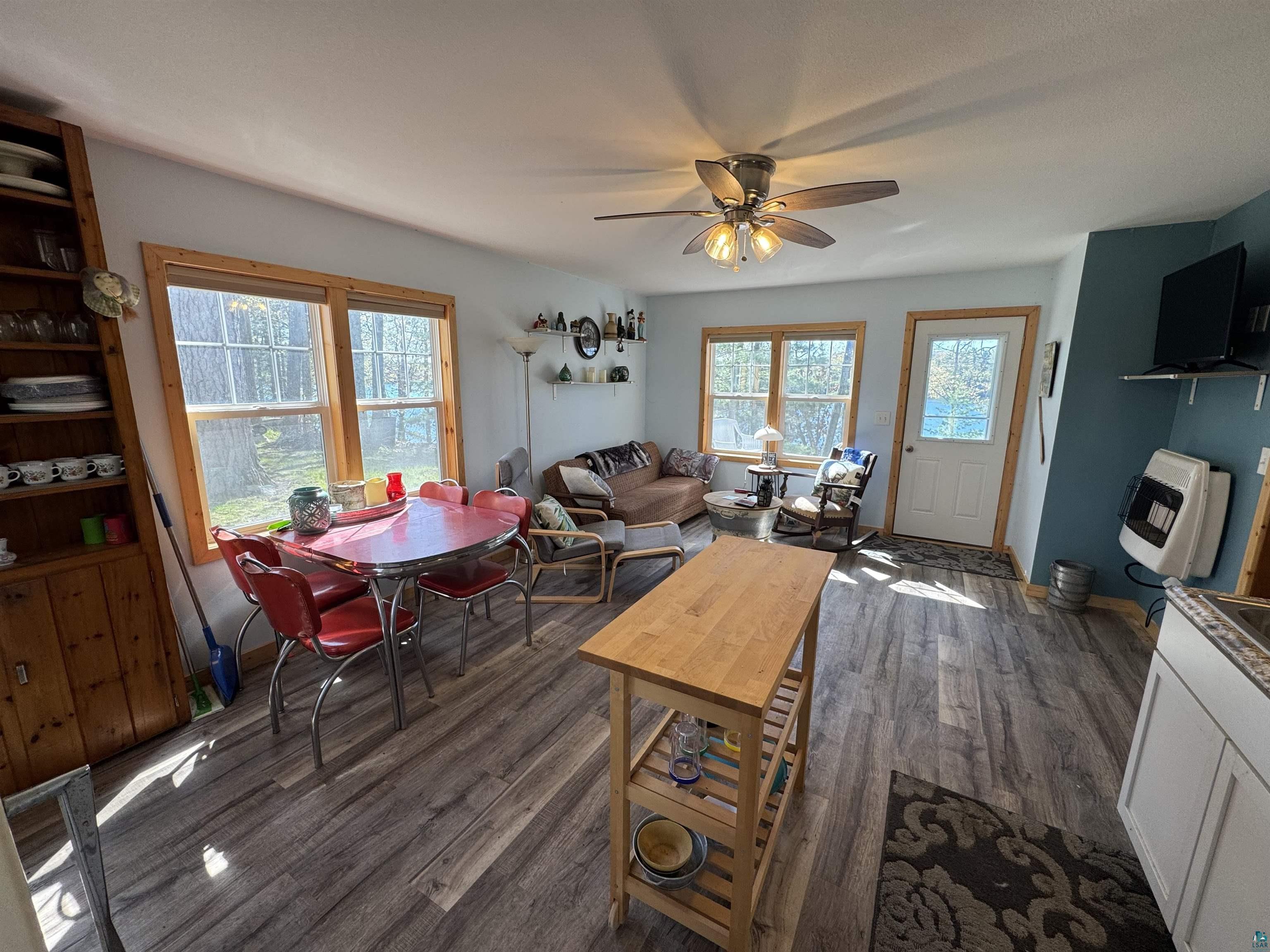 Image 2: Open-concept living area featuring wood-finish flooring, multiple windows with wooded exterior scenery, a ceiling fan with integrated lighting, and a built-in wooden shelving unit, Living Room