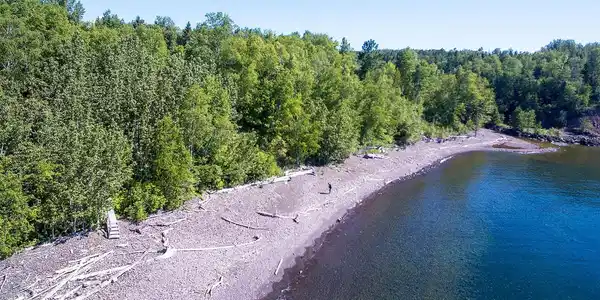 Image 1: Wooded shoreline property featuring a pebble beach, clear water, and mature trees, View
