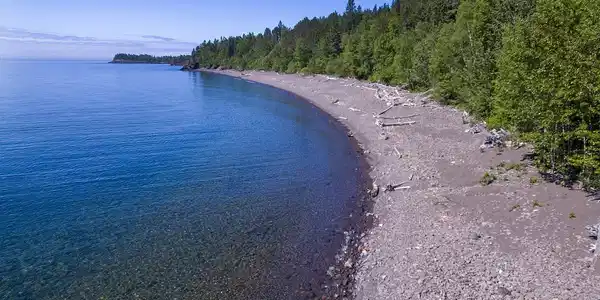 Image 2: Expansive shoreline featuring a pebble beach, clear blue water, and a dense treeline, View