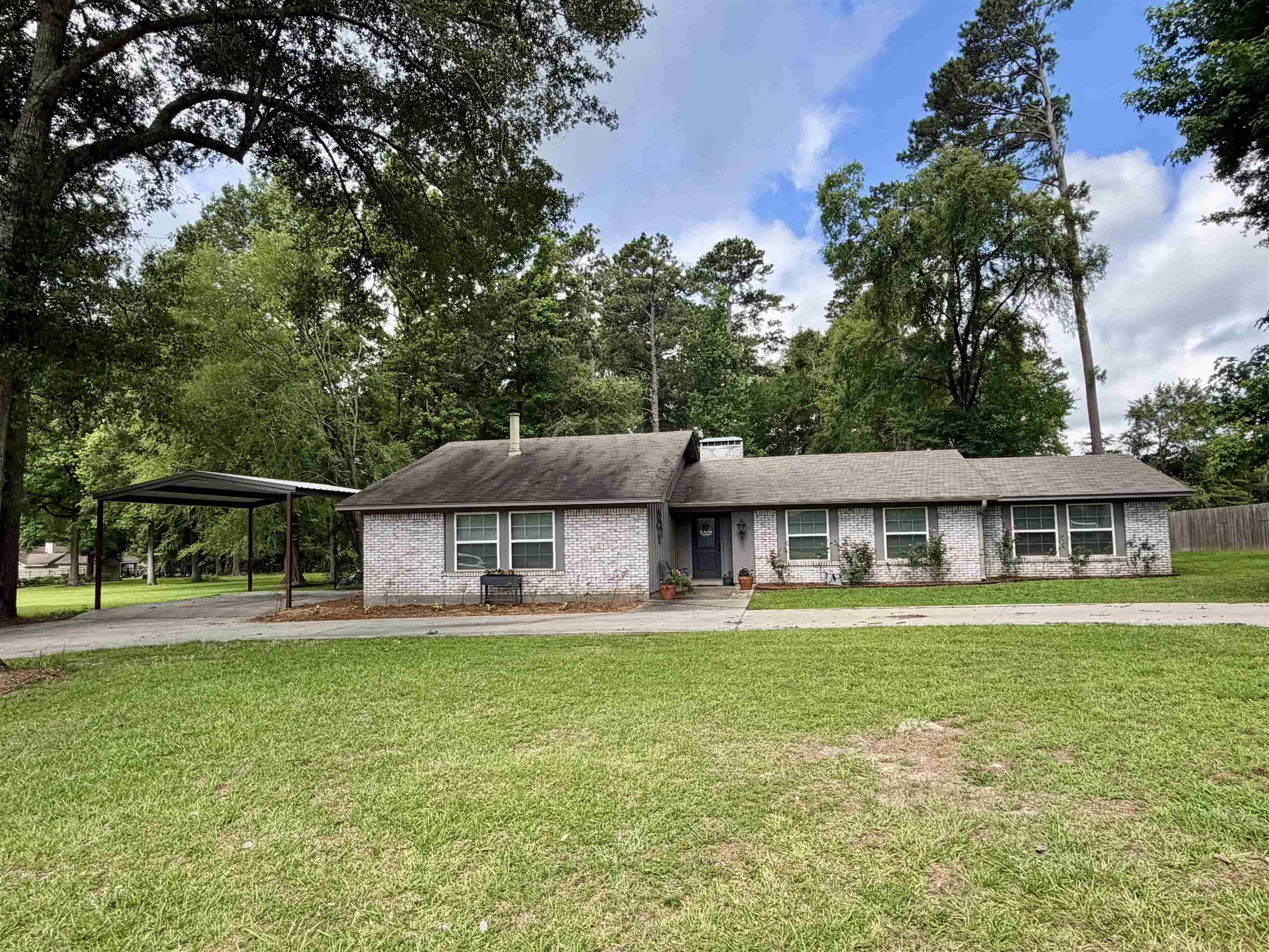 Image 1: Brick exterior with a gray-toned finish, featuring a carport, mature trees, a shingle roof, and a concrete driveway, Front Of Structure