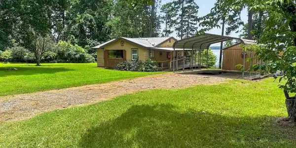 Image 3: Waterfront property featuring a single-story residence with light-colored siding, a metal carport, and an additional wood-sided structure, Front Of Structure