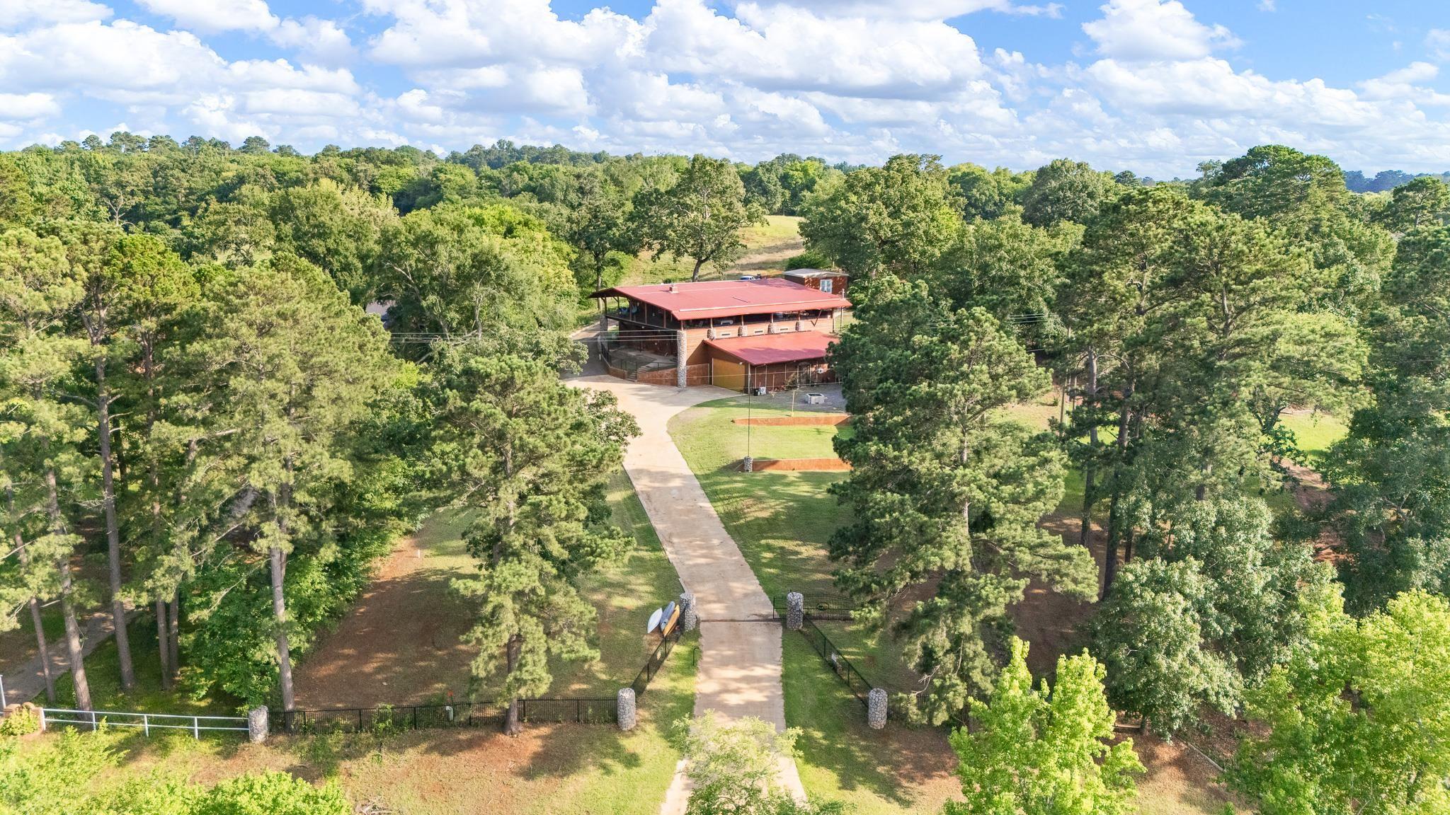 Image 1: Gated property with extensive tree coverage and a long driveway leading to a multi-level structure featuring a red roof and wood siding, Aerial View