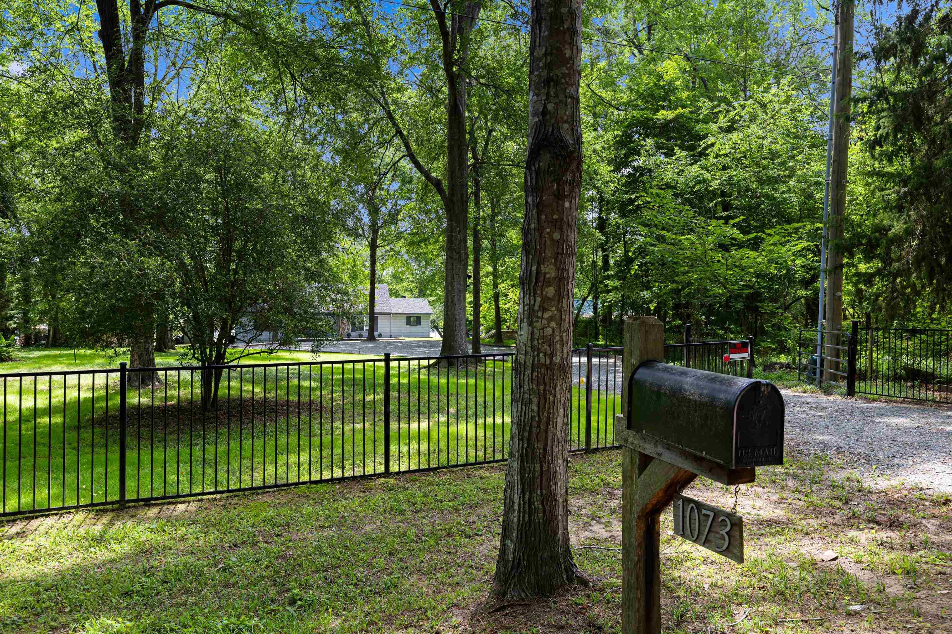 Image 1: Gated gravel driveway entrance featuring a black metal fence, mature trees, and a distant house with light-colored siding, Yard