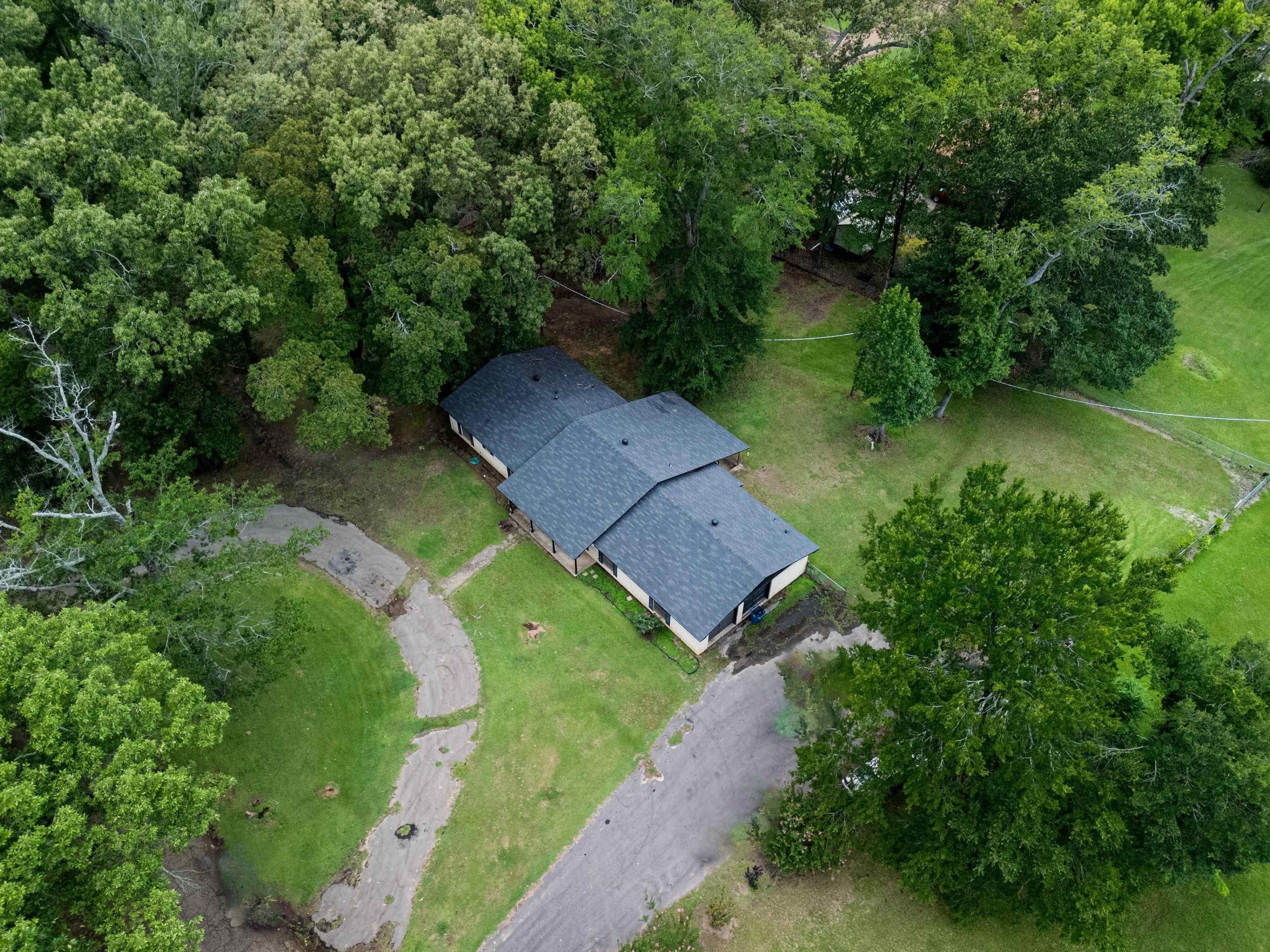 Image 3: Expansive property featuring a single-story residence with a dark shingle roof, surrounded by mature trees, a partially paved driveway, and substantial green space, Aerial View