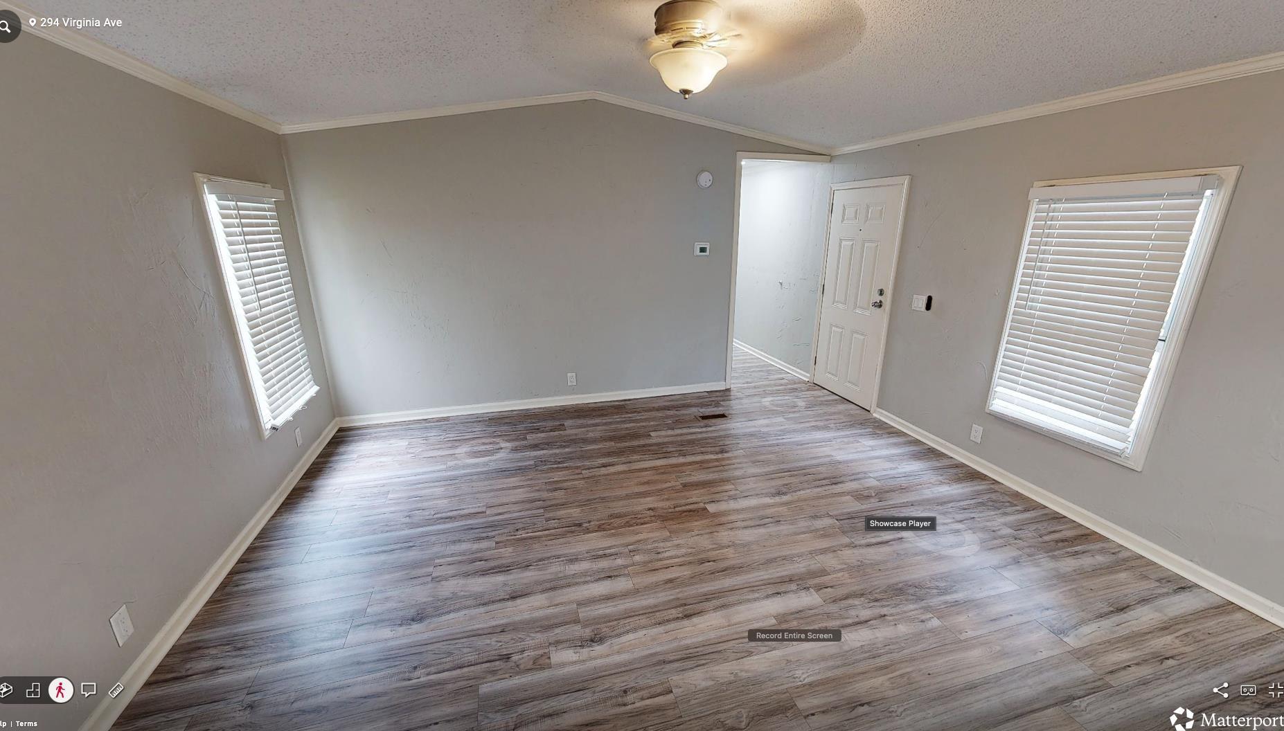 Image 3: Spacious room featuring wood-finish flooring, two windows with blinds, a white paneled door, and a ceiling-mounted light fixture, Living Room