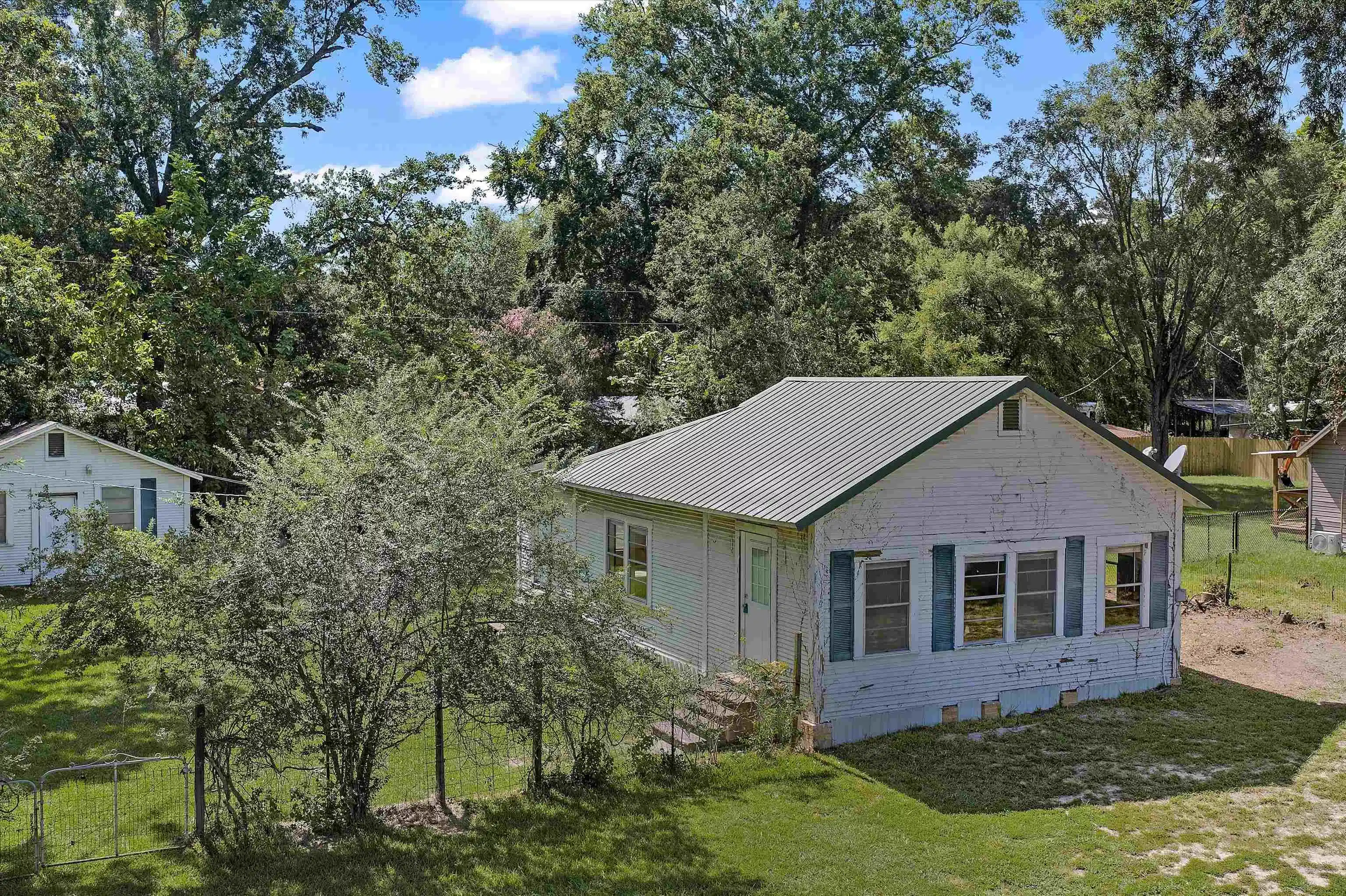 Image 0: Single-story residence featuring a metal roof, multiple windows with shutters, and a front yard with mature trees, Side Of Structure